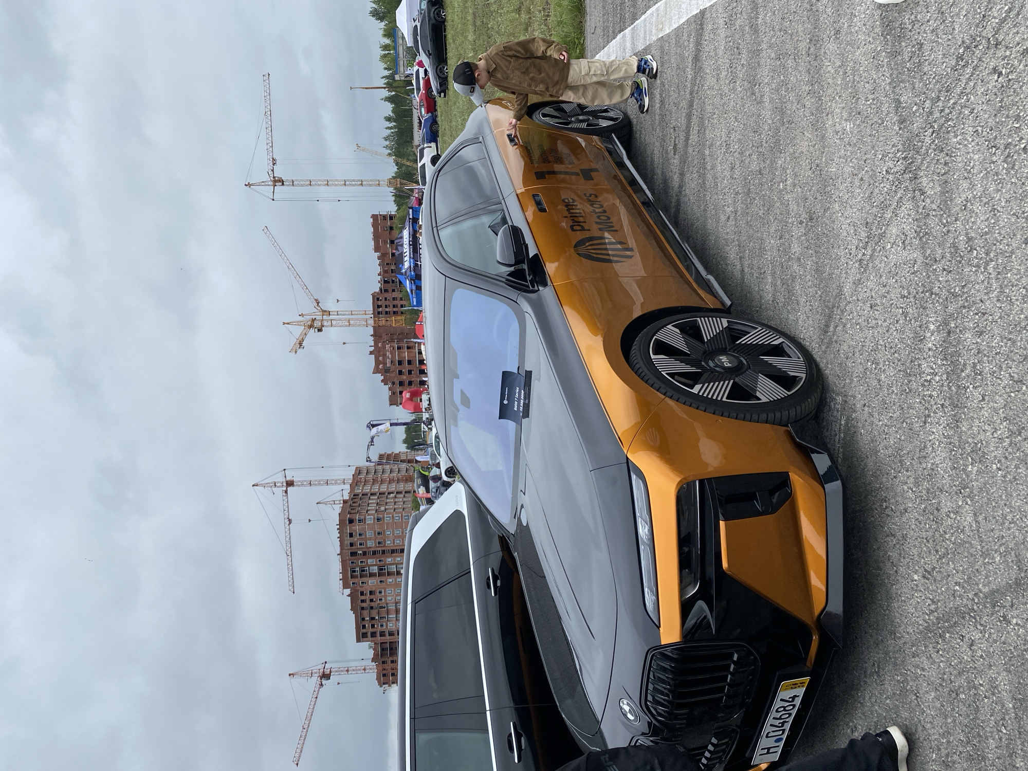 The image shows a two-tone BMW sedan, with a black hood and a bronze body, parked outdoors. A young boy in a brown jacket and khaki pants is standing by the car, touching the door handle. Behind the BMW, a black SUV is visible. In the background, there are several construction cranes and unfinished buildings, suggesting a construction site or a developing urban area. Further in the distance, other cars and tents can be seen. The sky is overcast. The license plate of the BMW appears to be H.04684. The scene is likely a car show or an outdoor event near a construction zone. There is no direct visual information in the image to confirm the specific location within the city of Perm, Russia.