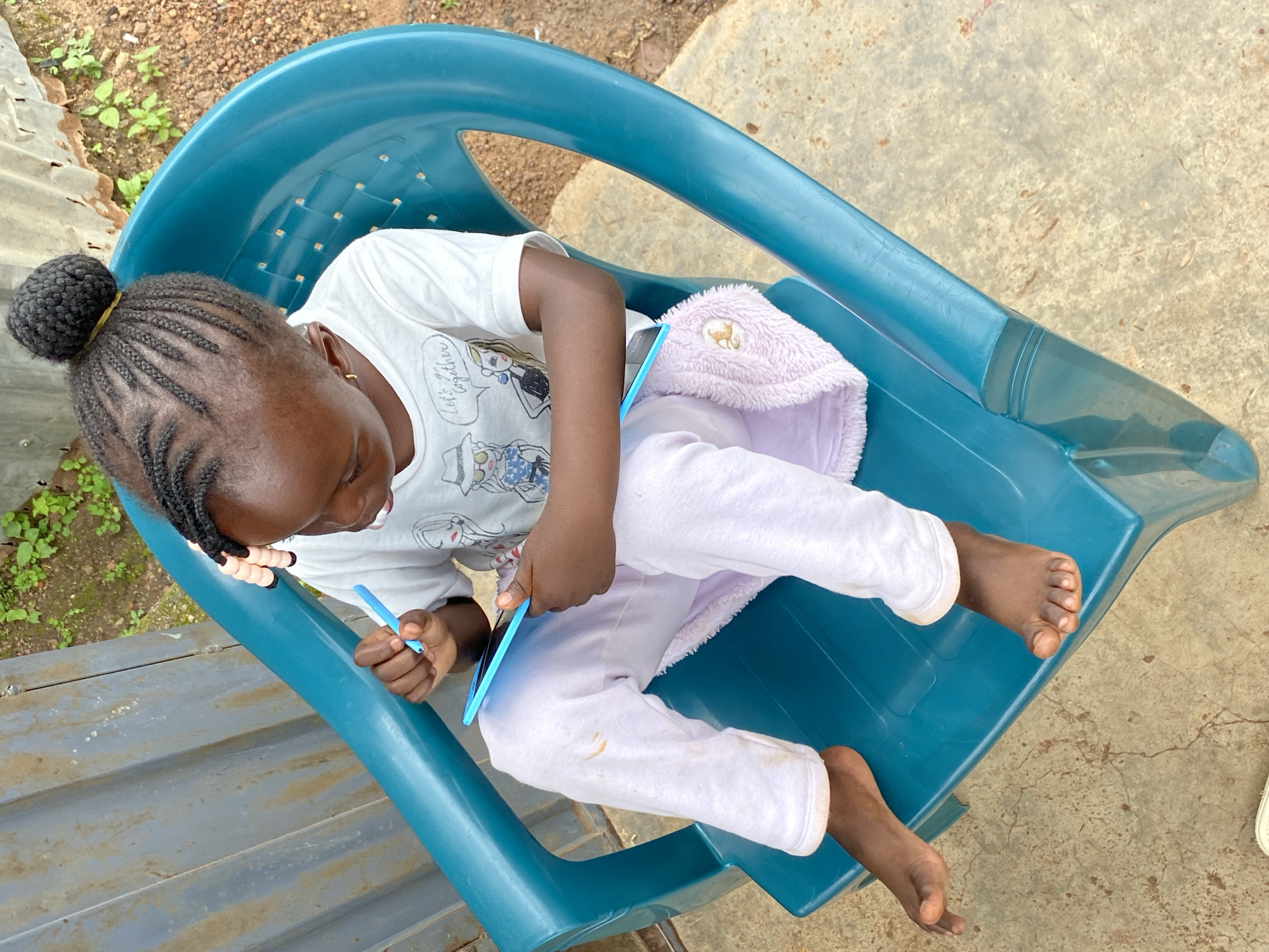 A young Black girl with braided hair tied into a bun is sitting on a teal plastic chair outdoors. She is wearing a white t-shirt with cartoon characters and text that appears to say "Let's Get Together" and light pink sweatpants with a fluffy pink pouch attached. The girl is holding a blue stylus and appears to be interacting with a blue tablet resting on her lap. Her feet are bare, and her toes are visible. The chair is on a cracked concrete surface, and behind it is a corrugated metal fence or wall, with some greenery visible beyond. The ground around the chair is dirt and pebbles. The scene suggests a casual, outdoor setting, possibly a yard or courtyard.