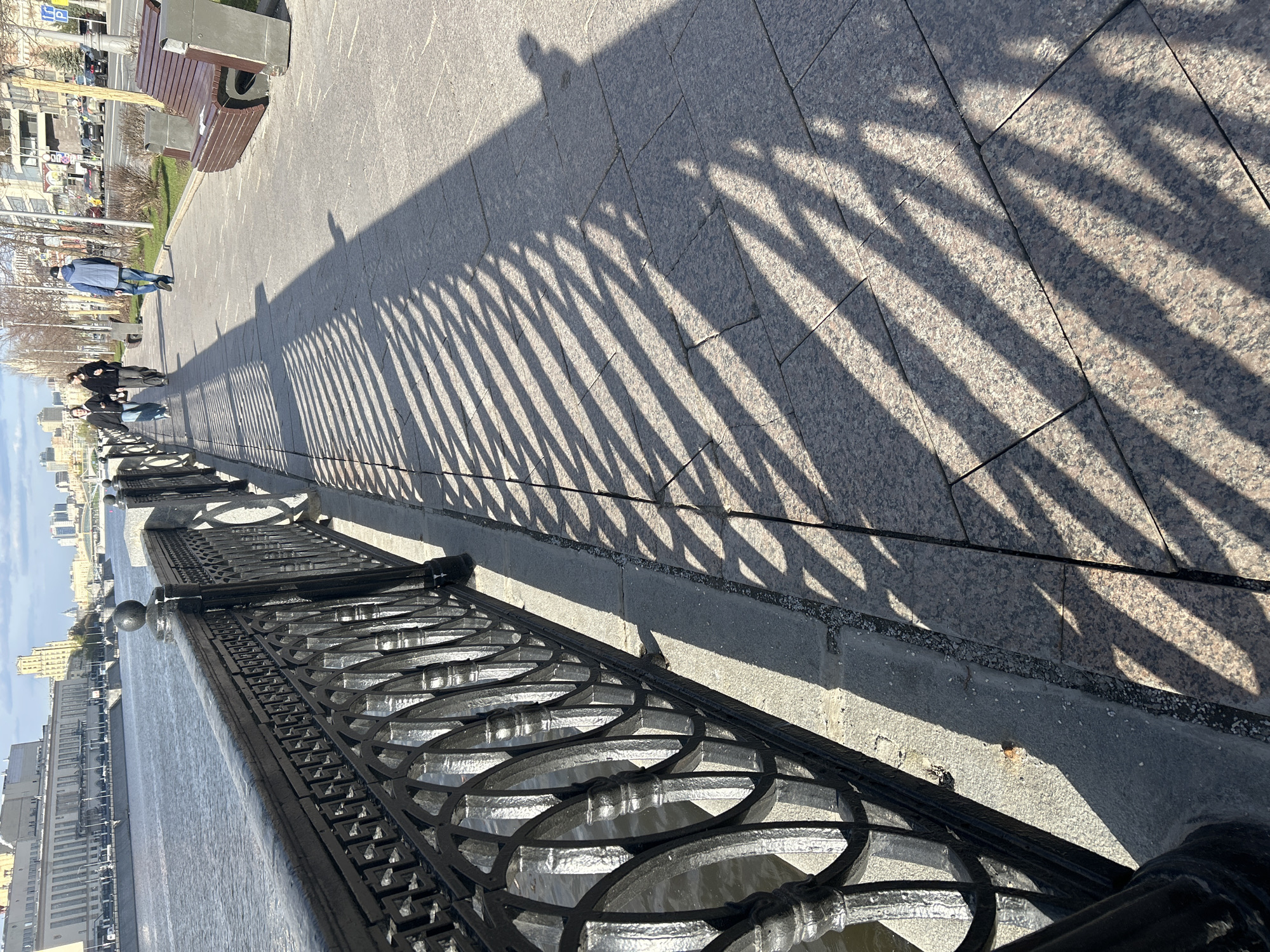 A ground-level, eye-level, medium-distance shot depicts a city street scene next to a river, possibly in Moscow, Russia. A black wrought iron fence runs along the left side of the image, alongside the water. The fence's design includes circular elements.
A paved walkway extends along the right side of the frame, composed of large, rectangular stones. The sunlight casts a striped shadow from the fence onto the walkway. A few individuals are walking along the path. In the background, there are buildings, trees, and street furniture, including a bench. The light suggests it is a sunny day.