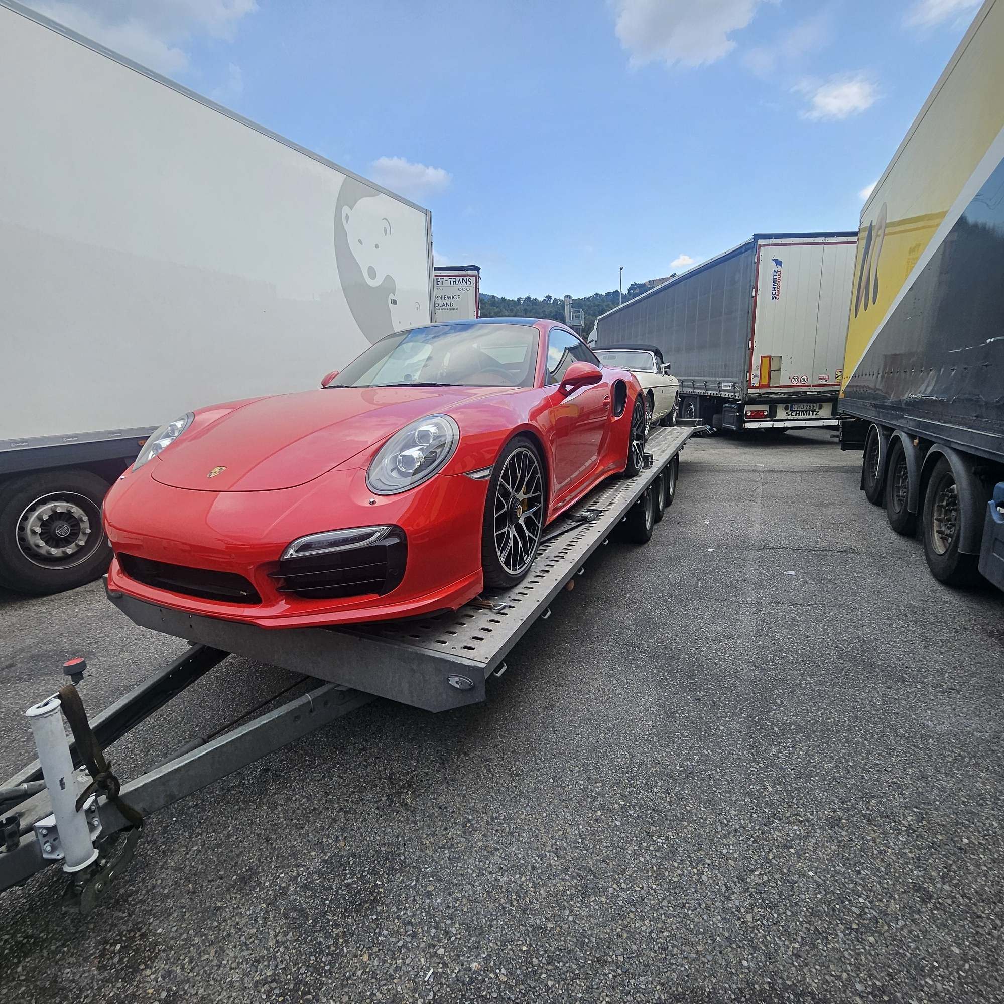 A bright red Porsche 911 is parked on a car carrier trailer, which is on an asphalt surface, likely a parking lot or rest stop. The car is positioned at an angle, with its front end facing left. The trailer has multiple wheels and appears to be in a state of transport or loading. In the background, several large trucks are parked, including a white trailer with a circular logo, a white trailer with "SCHMITZ" branding, and a yellow trailer with black graphics. The sky above is blue with scattered white clouds. The overall impression is one of a commercial vehicle stop or transport hub. The scene is located in La Jonquera, Spain. There are no people visible in the image.