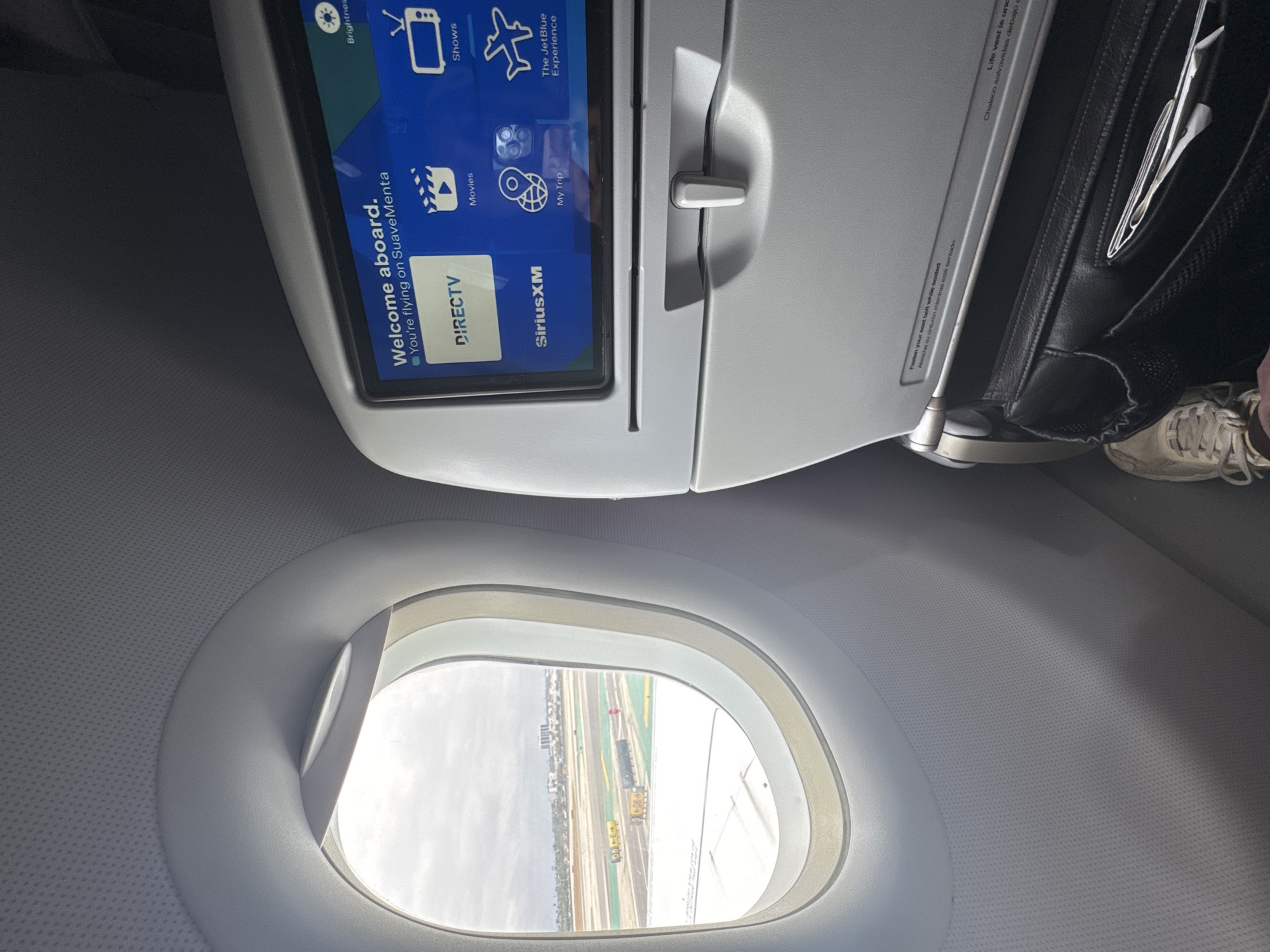 An interior view of an airplane cabin shows a window on the left and a seatback entertainment screen on the right. The window looks out onto an airport tarmac with a cloudy sky overhead. Several yellow signs and some parked vehicles are visible on the tarmac. The entertainment screen displays "Welcome aboard. You're flying on SuaveMenta" and icons for DIRECTV, Movies, Shows, SiriusXM, My Trip, and The JetBlue Experience. Below the screen is a tray table latch and a seat pocket. A person's foot wearing a light-colored sneaker is visible in the lower right corner of the image. The image appears to have been taken at Los Angeles International Airport (LAX), given the visible infrastructure of an airport and the general appearance of the surrounding environment.