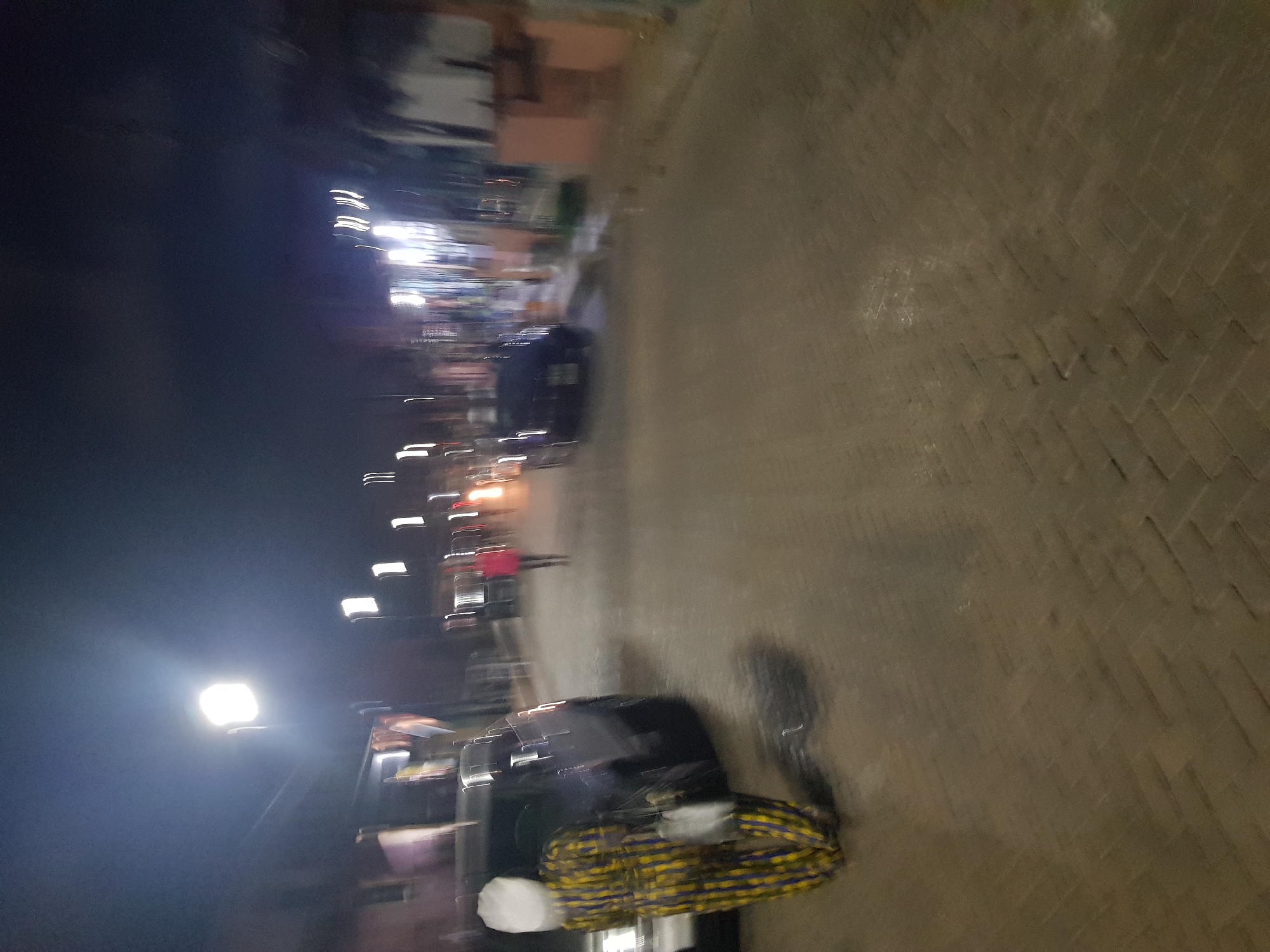 At night in Shangisha, Nigeria, a blurred image depicts a street scene. The brick-paved road occupies much of the foreground. Two dark-colored cars are visible, one closer to the viewer with what appears to be a large, possibly woven object on its roof. Streetlights and illuminated buildings are seen along the left side of the image, further down the road. A figure can be seen on the side of the street.