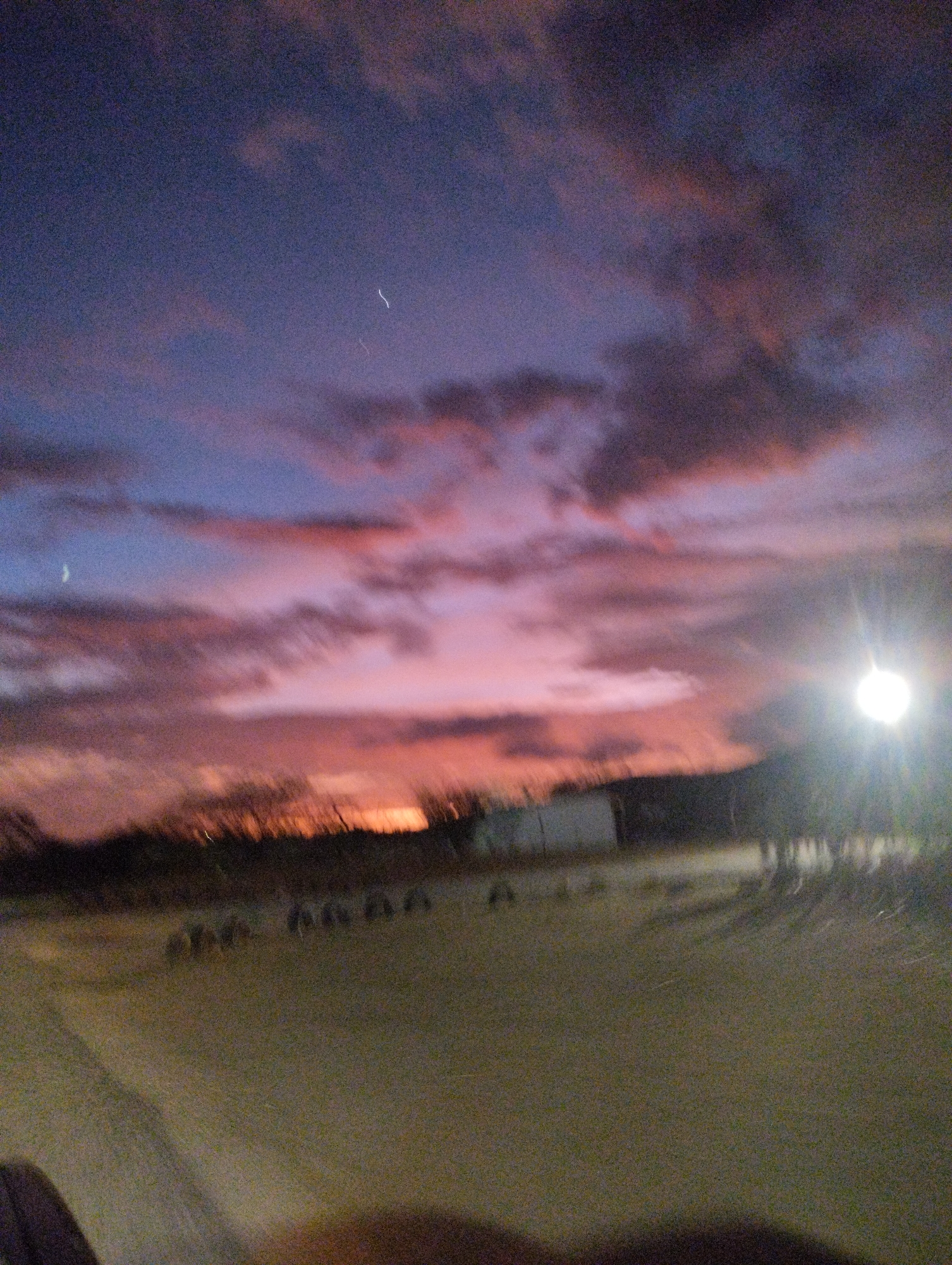 The image shows a blurred outdoor scene at dusk. The sky is filled with clouds tinged with pink and purple hues, indicating a sunset or sunrise. A bright light source, possibly a streetlamp or a powerful bulb, is visible on the right side of the frame, creating a glare. In the background, there are indistinct shapes that could be buildings or structures, and a line of trees or vegetation forms a silhouette against the colorful sky. The foreground is a flat, textured surface, possibly pavement or dirt. There are some faint, elongated shapes in the middle ground that are too blurry to identify precisely, but they could be abstract representations of people or objects. The overall impression is one of low light conditions and motion blur, suggesting the photo was taken at night or during low light with a moving camera. The location mentioned is La Guardia, Venezuela.