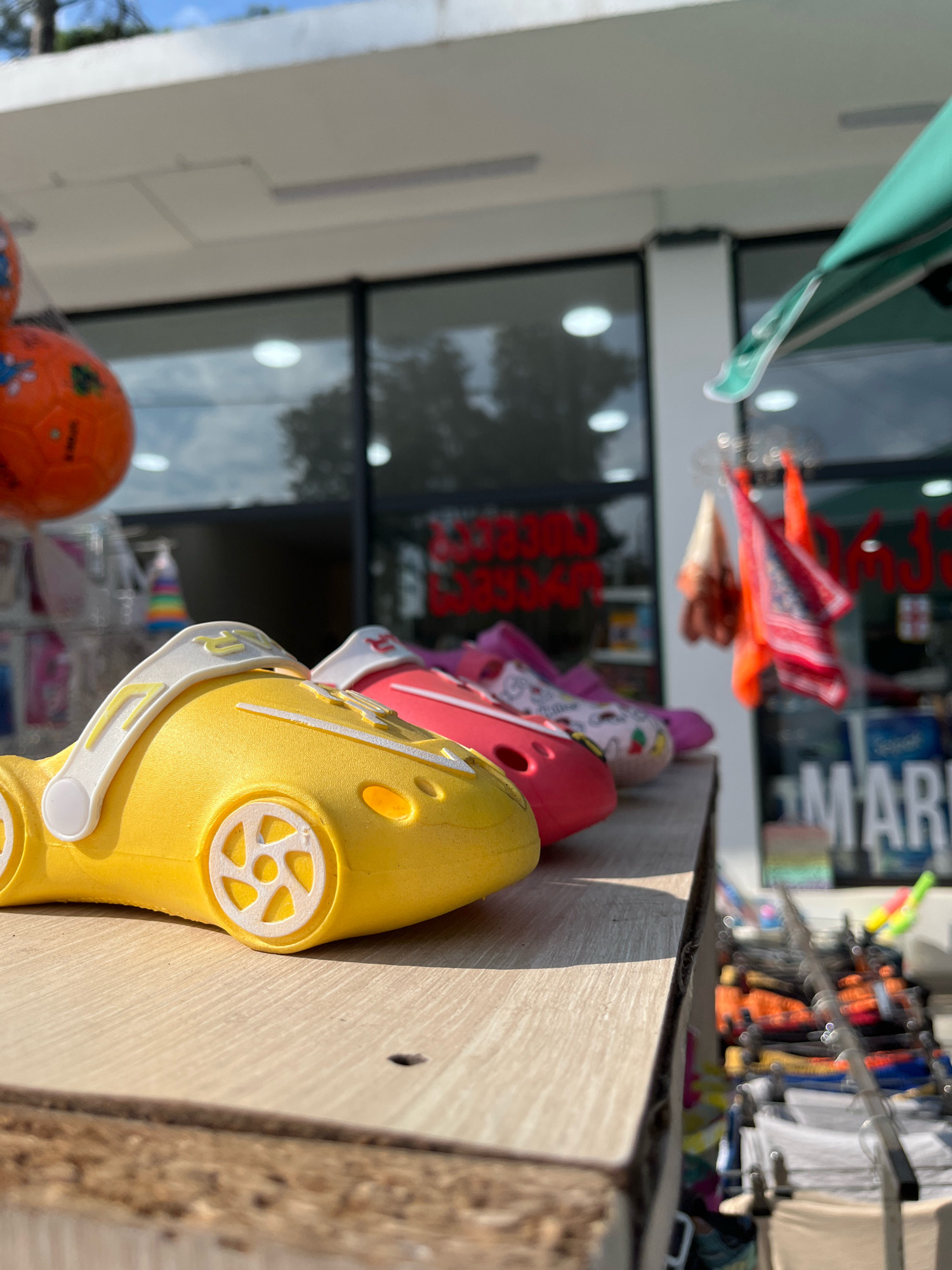 The image shows a close-up view of several colorful children's character clogs lined up on a wooden shelf. The clogs are shaped like cars. The closest clog is yellow and features white wheels with a five-spoke design. Behind it, there is a pink clog with a white strap adorned with colorful graphics of animals and hearts, and further back, a red clog. The background is blurred but appears to be an outdoor market or storefront in Ureki, Georgia. Through the glass of the storefront, various items are visible, including what appear to be colorful towels or scarves hanging, and racks of clothing or merchandise. A green awning is visible at the top right of the image. The sky above is bright and clear, suggesting a sunny day. The overall impression is of a vibrant and casual retail environment, likely catering to tourists or local families.