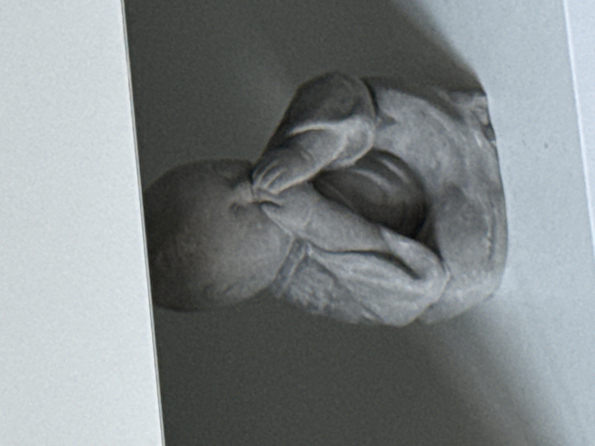 A close-up, eye-level shot shows a small, grey, stone statue of a baby with its hands covering its mouth, possibly in a gesture of silence or surprise. The statue is positioned on a flat, light grey surface, likely a shelf or table. Above the statue, a darker grey surface is visible, creating a shadowed overhang. The background is a muted, dark grey. The image does not provide any information about a specific location in Stara Zagora, Bulgaria, or any actions, interactions, or time of day.