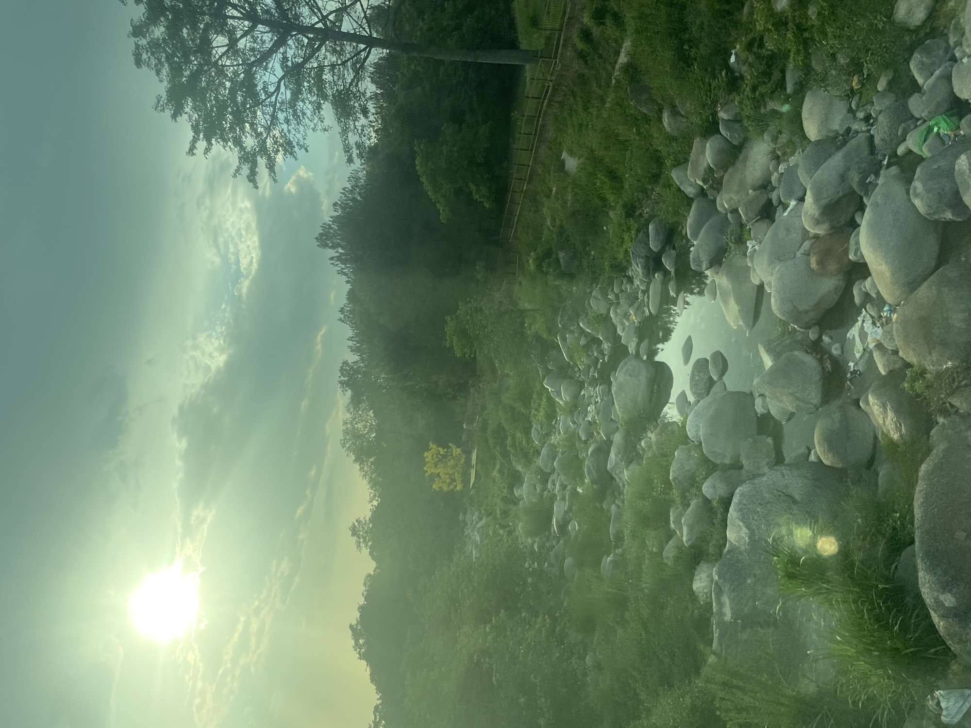 The image depicts a rocky riverbed in Chetru, India, bathed in sunlight. The river itself is shallow, with numerous large, rounded rocks scattered along its course. Vegetation, primarily grass and trees, grows along the banks. A wooden fence runs along the upper part of the riverbank on the far side. The sky is overcast with the sun visible through the clouds.