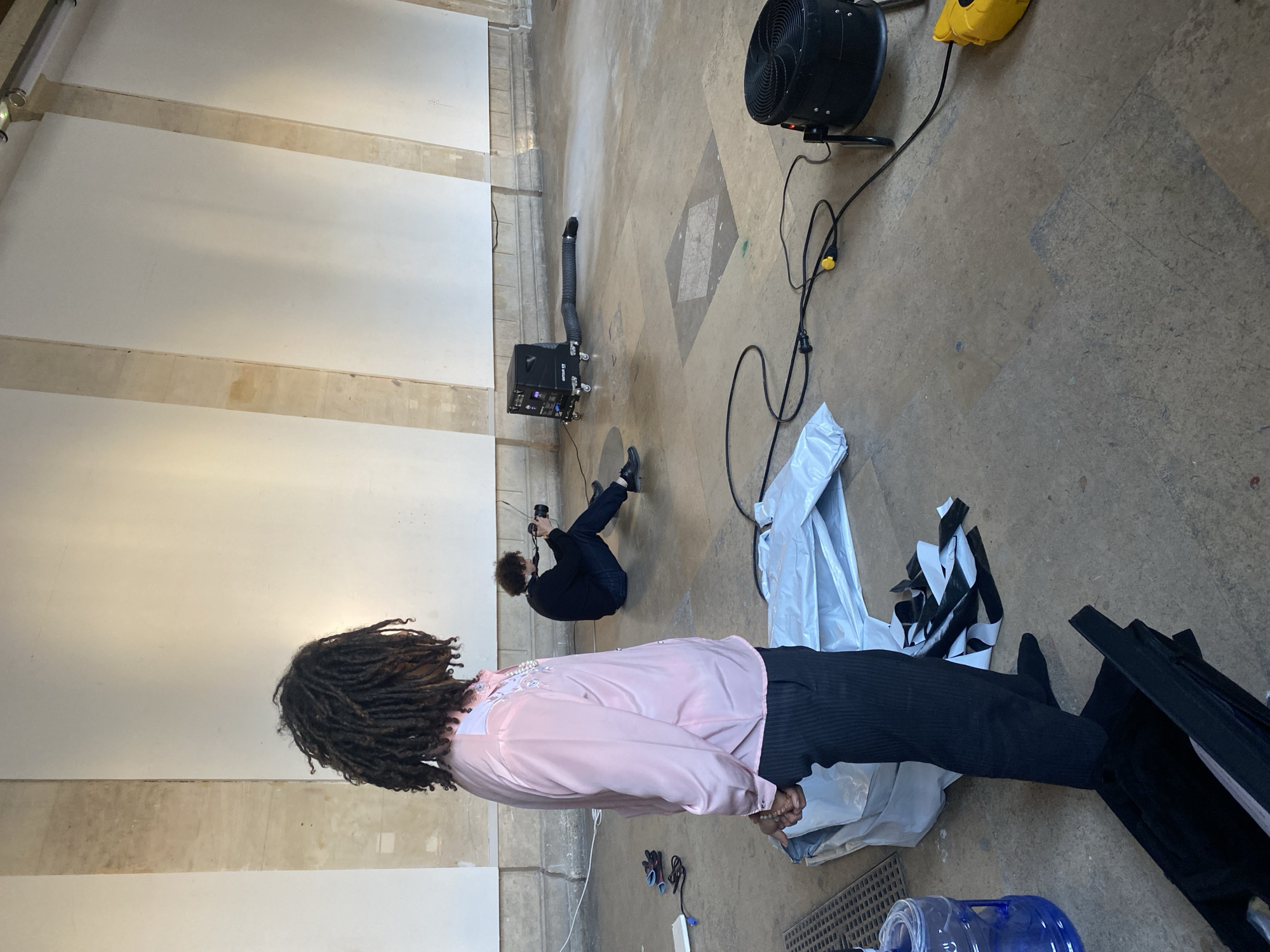The image depicts an indoor scene that appears to be a photoshoot or video shoot in progress.
**Subjects:** There are two individuals present. In the foreground, with their back to the viewer, stands a person with long dark dreadlocks. They are wearing a pink satin shirt with white embellishments on the shoulders and dark, possibly pinstriped, trousers. Their hands are clasped in front of them, holding what looks like a string of beads or a small object. In the midground, another person with curly dark hair is crouched on the floor, operating a black digital camera, seemingly focused on the person in the foreground.
**Scene/Setting:** The setting is a spacious, minimally decorated indoor location, possibly a studio or a renovated industrial space. It features a light-colored concrete or tiled floor with subtle darker geometric patterns. The high walls are painted white, punctuated by several horizontal, light-colored structural beams or supports. A tubular light fixture is visible on the upper left wall.
**Activity or Event:** The primary activity is a professional visual media production, with one person posing as the subject and another behind the camera.
**Notable details:** Various pieces of equipment are scattered on the floor: a large black industrial-style fan with a circular guard, a black box-like machine with a flexible black hose extending from it (likely a fog or haze machine), and a yellow power distribution unit connected by several black cables. Other items include a large clear blue water bottle, a pair of black gloves, and some crumpled white and black material. The indoor setting means the time of day is not apparent. There are no specific visual cues within the image to indicate the provided location context of Paris, France.
**Visible text:** The word "HAZER" is visible on the side of the black box machine with the hose.