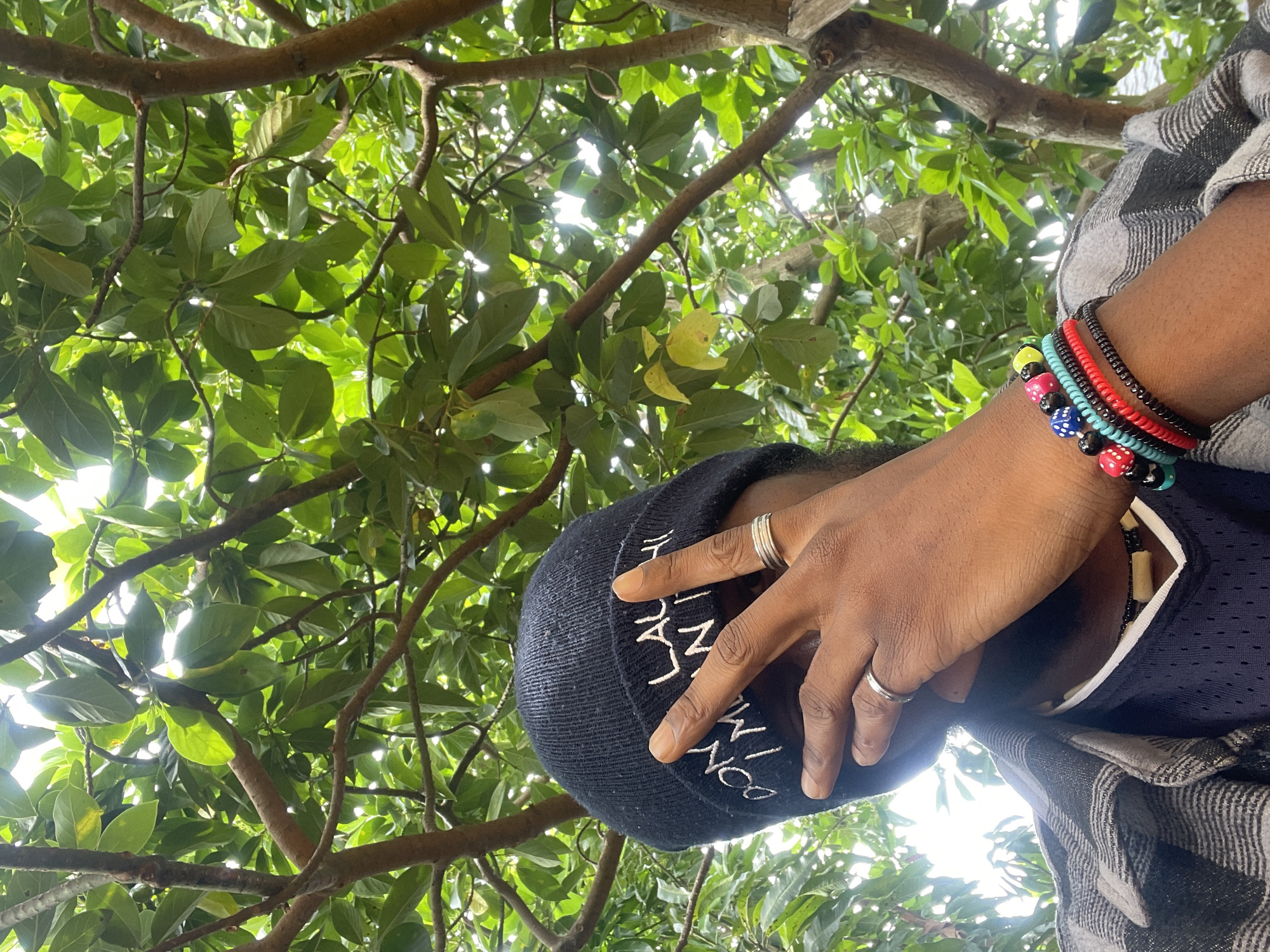 A low-angle medium shot depicts a person standing beneath a tree, likely in Jos, Nigeria, based on the background foliage and skin tone. The person is wearing a dark blue beanie with white lettering, a dark blue shirt with white trim, and a gray-and-white checkered overshirt. Their right hand, adorned with two silver rings, is raised towards the beanie. Their left wrist features a stack of colorful bracelets, including one with dice-patterned beads. The background consists of dense green foliage and branches, with sunlight filtering through the leaves.