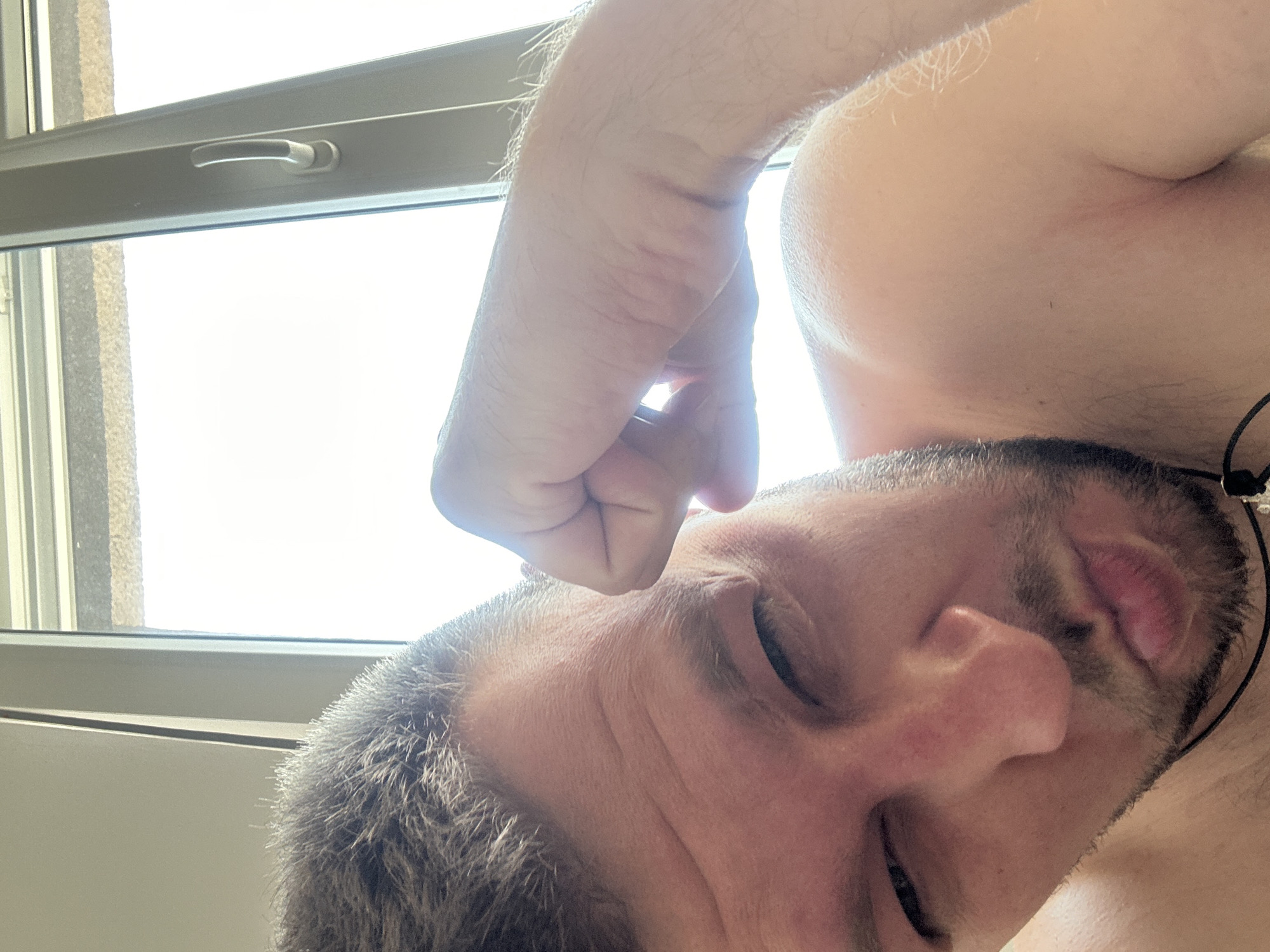 A close-up, low-angle selfie shows a man with short, dark hair and stubble, looking towards the camera with his right eye partially closed and his lips pursed. His right arm is raised, with his fist resting near his temple. He appears to be indoors, with a window and a bright sky visible in the background. The man is shirtless, and a dark cord necklace is visible around his neck. The scene is lit by natural light coming from the window. There is no explicit indication of the location in Zvezdara Urban Municipality, Serbia.