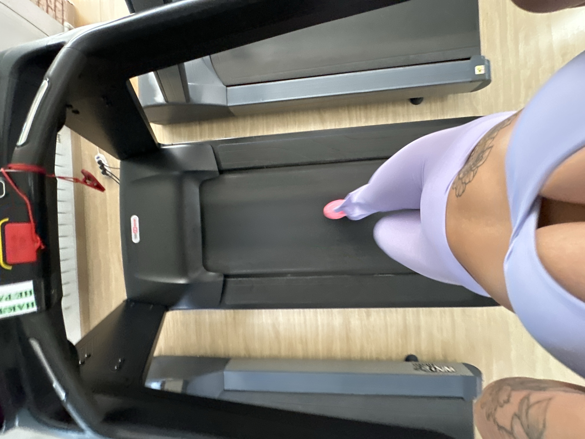 The image shows a close-up, angled view of a person on a treadmill in a gym. The person is wearing light purple athletic wear and has a tattoo on their lower back. Their right foot, clad in a pink sneaker, is visible on the moving belt of the treadmill. The treadmill itself is black with silver accents. In the background, another treadmill is partially visible. The setting appears to be an indoor gym with a light-colored wooden floor. The lighting suggests it is daytime. There is text visible on a white and green sticker on the left side of the treadmill, partially obscured, reading "HEALTHE PA..." and some other unreadable characters. Another sticker with red and white text is visible on the treadmill console. The perspective is from above and to the side of the person on the treadmill, focusing on their lower body and the exercise equipment.