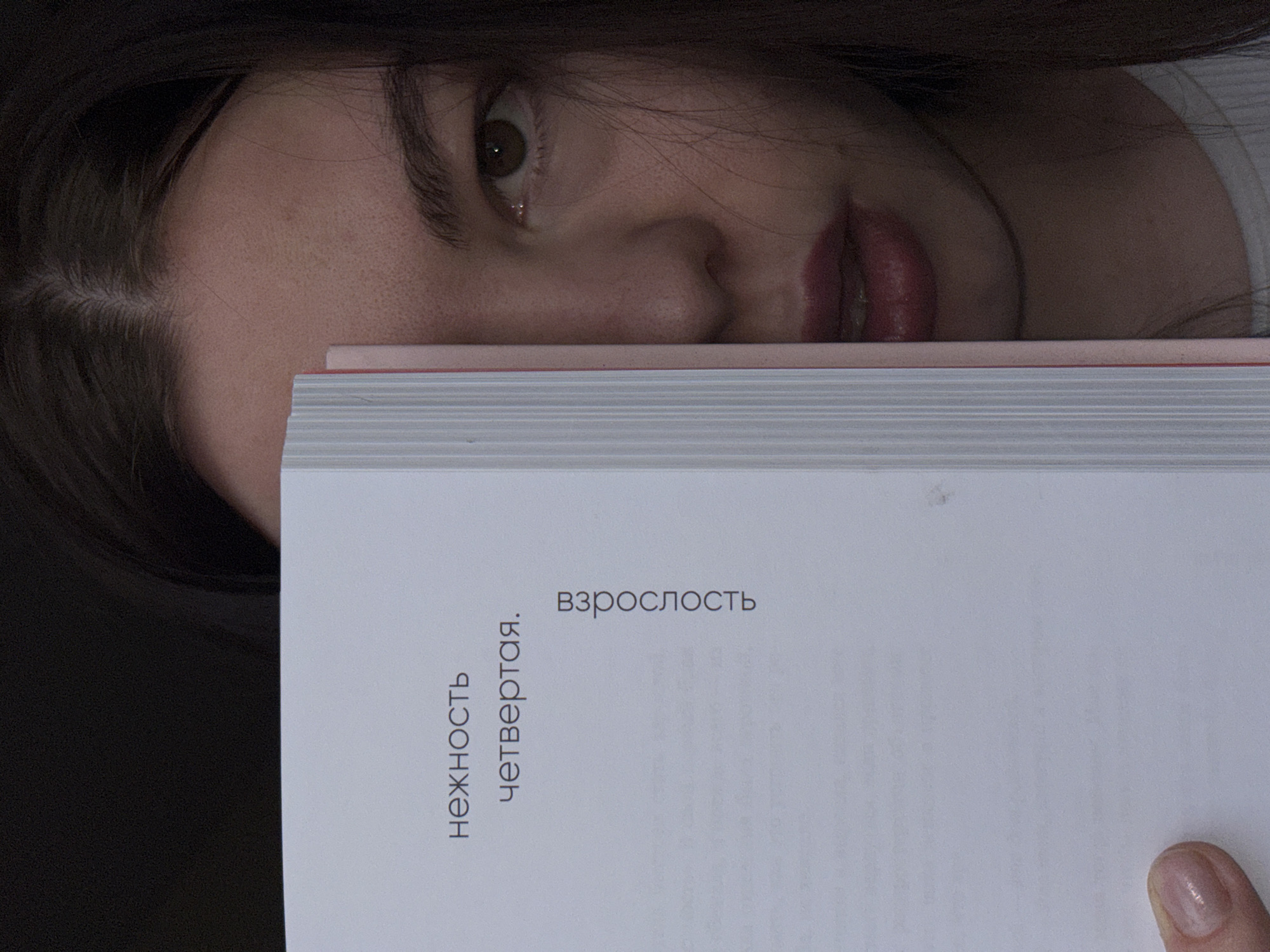 A woman is partially visible behind a book with Russian text. Her face, including an eye, nose, and lips, is visible above the book. The book is open to a page with the words "нежность" and "взрослость" printed on it. A thumb with a nail polished in a neutral color is visible at the bottom right corner of the book. The background is dark. The image does not contain enough information to determine the location in Perm, Russia.