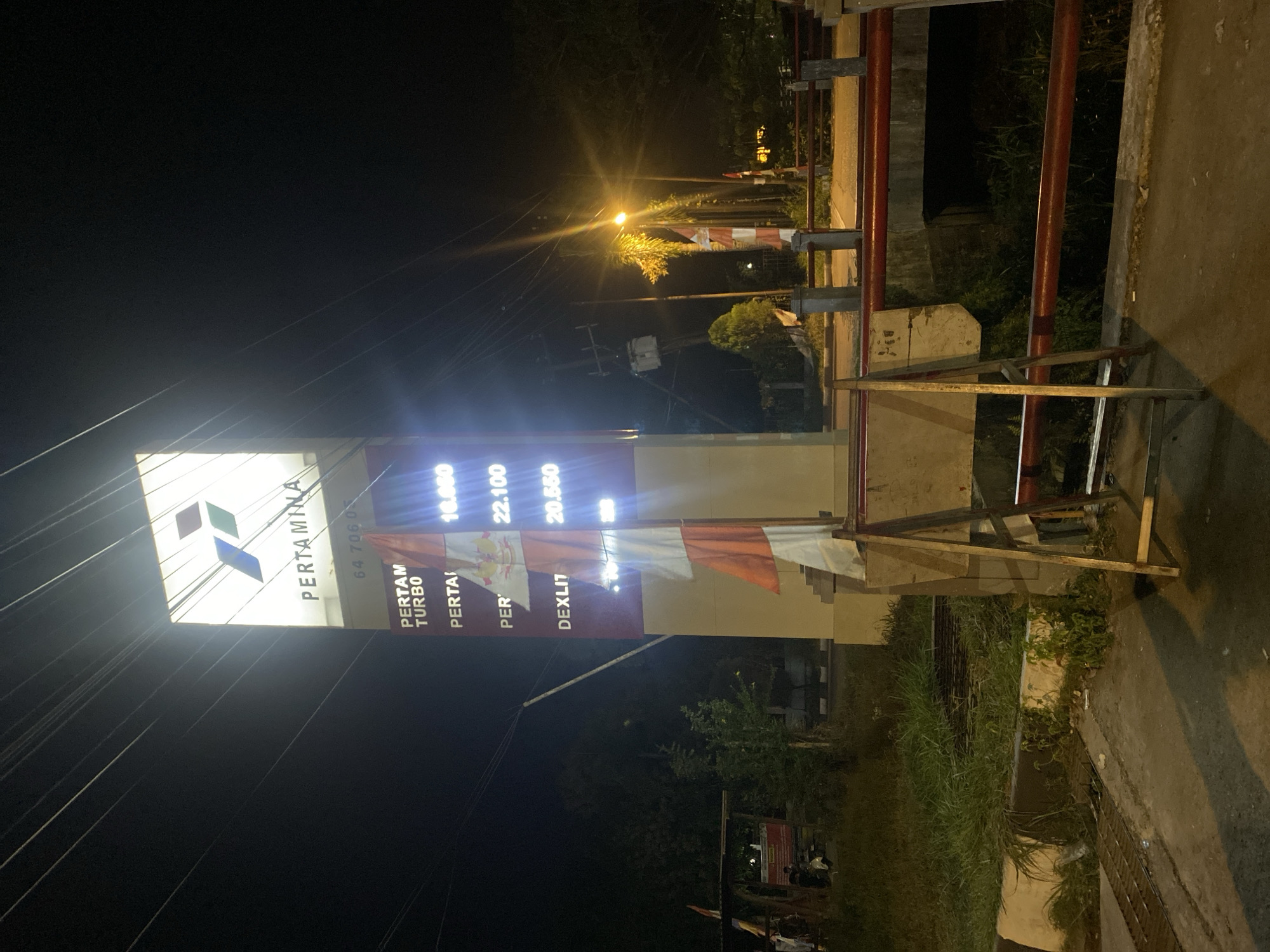 The image shows a Pertamina gas station sign at night in Gambut Barat, Indonesia. The illuminated sign displays the Pertamina logo and brand name at the top, followed by a phone number "64 706 07". Below this, a price list indicates "PERTAM TURBO" at 10.000, "PERTAI" at 22.100, and "DEXIT" at 20.550. Several Indonesian flags, red and white, are attached to the signpost. In the foreground, a makeshift wooden A-frame structure is present. The background reveals a street with power lines, trees, and a bridge with red railings. The overall scene is dimly lit, with the gas station sign being the primary light source.