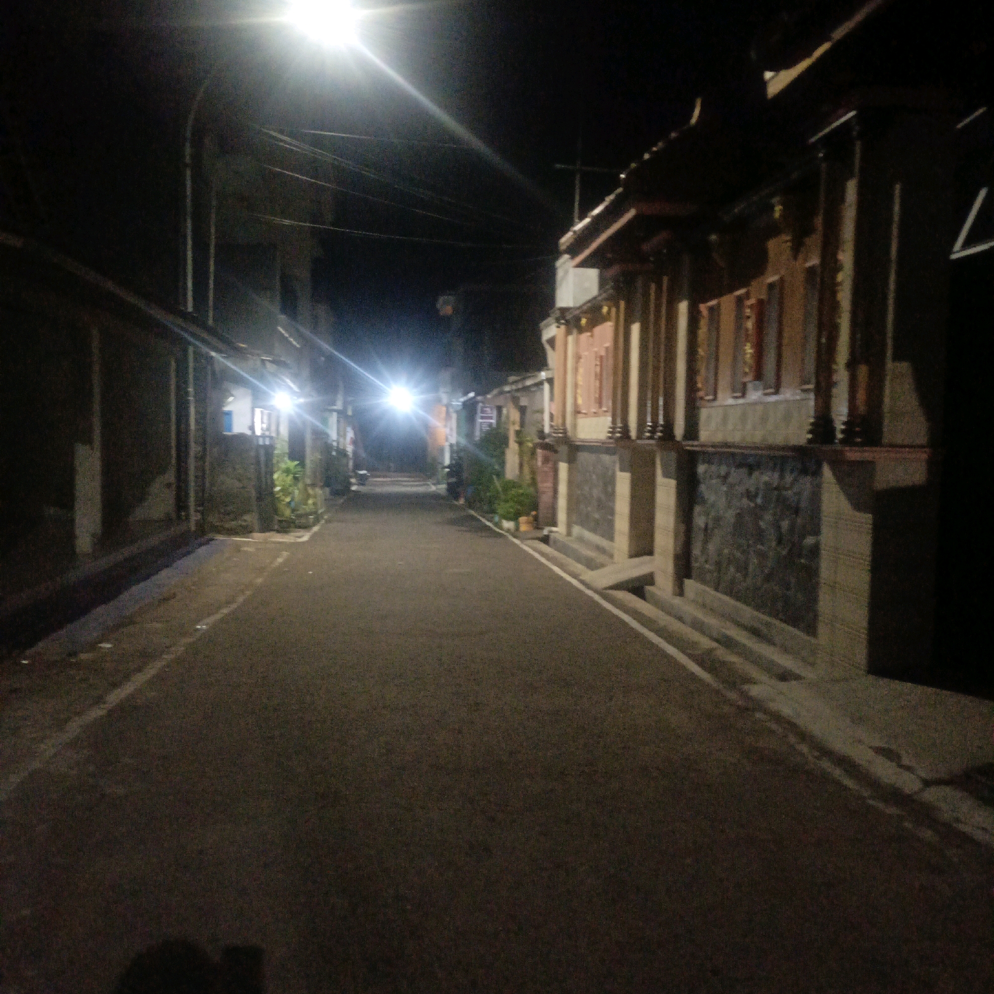 A nighttime street scene in Salatiga, Indonesia, shows an asphalt road with white lines marking its edges. On the right side of the road, a building with a dark roof and ornate pillars lines the street. The building's facade features stone and wood elements, with several windows or openings visible. Some greenery is present in front of the building. On the left side of the road, there are more buildings with stone walls, and some vegetation. Streetlights illuminate the scene, casting bright light and creating lens flares in the dark sky. The overall impression is of a quiet, residential street at night.