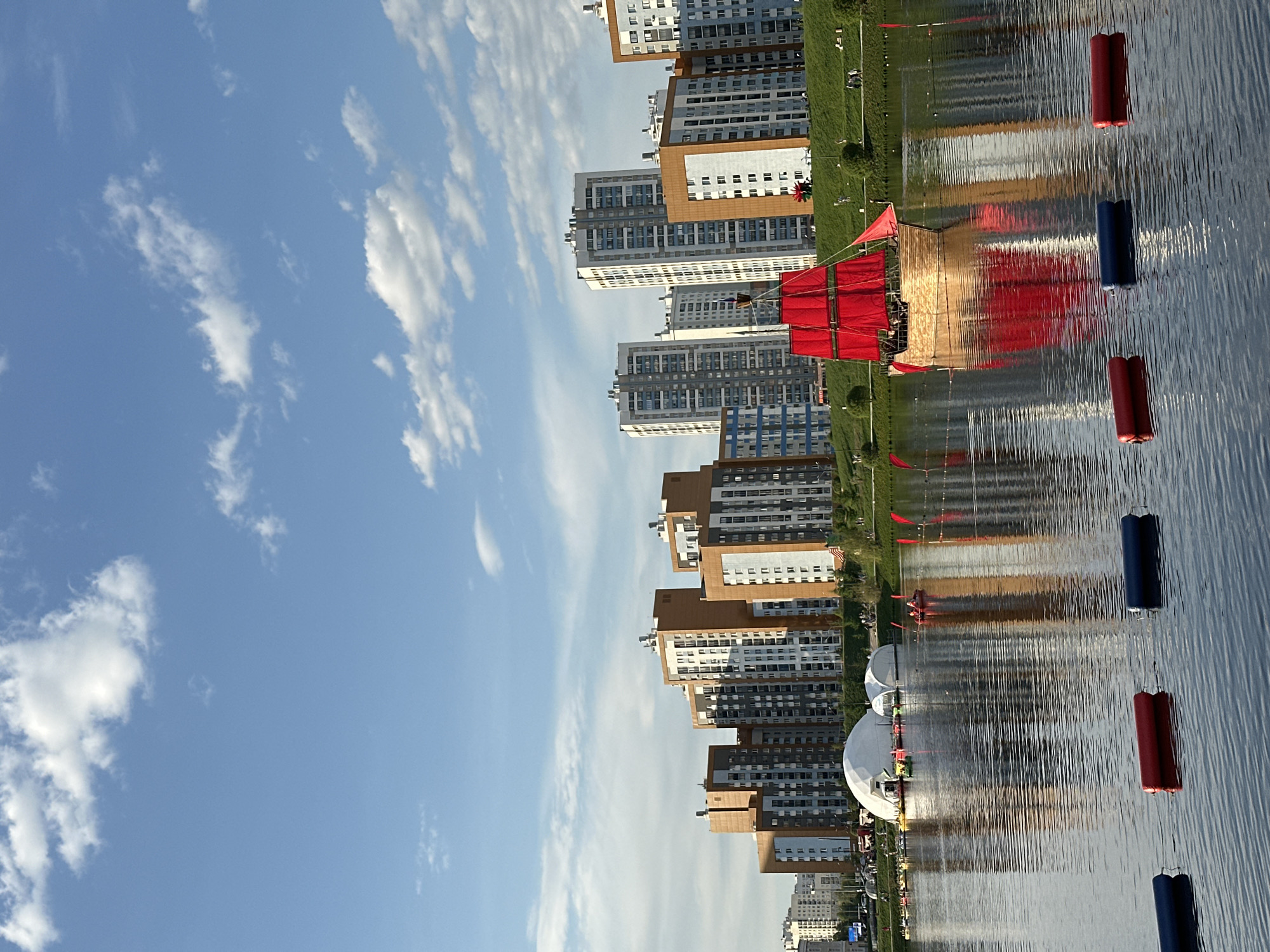 The image shows a view of the Iset River in Yekaterinburg, Russia. Several high-rise apartment buildings line the shore, their reflections visible in the water. Along the river, there are large white dome structures, possibly event venues. Red and blue cylindrical buoys float on the river's surface. A structure with red canopies is also present on the river. The sky is blue with scattered white clouds.