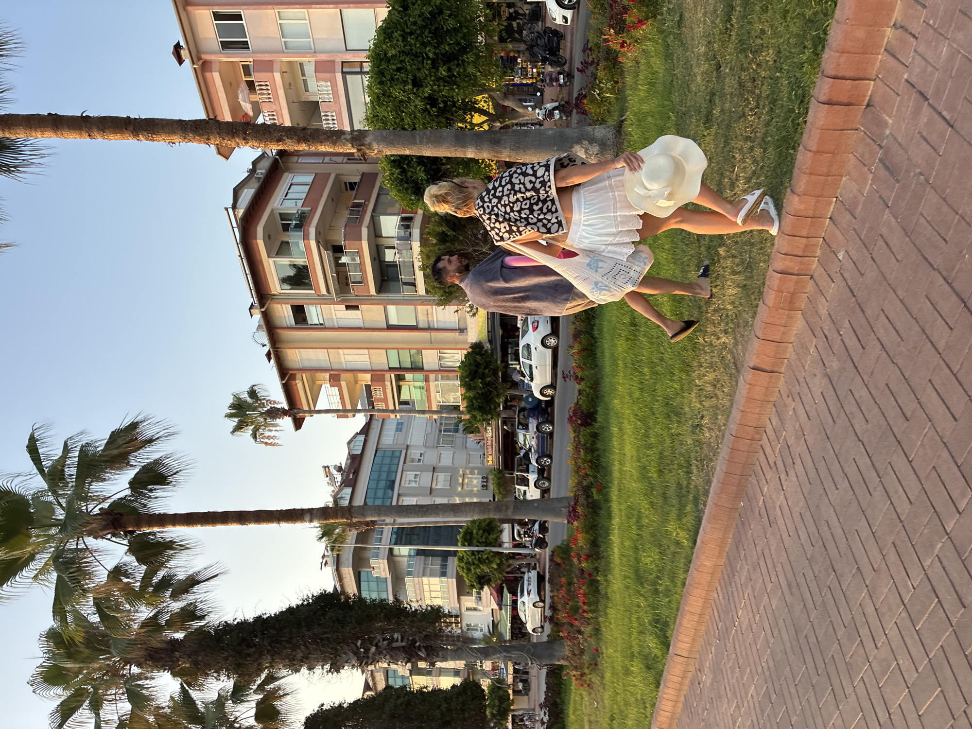 This image depicts a scene on a street in Alanya, Turkey, likely near the Cleopatra Beach area, given the palm trees and pedestrian walkway. The sun is low in the sky, casting long shadows and a warm glow.
In the foreground, a paved walkway made of reddish-brown rectangular bricks runs along the bottom of the frame. A low, brick border separates the walkway from a meticulously maintained strip of vibrant green grass. Beyond the grass, a paved road is visible, with several vehicles parked or driving on it. These include a white sedan, a few white open-top vehicles resembling golf carts or small buggies, a dark blue open-top vehicle, and some motorcycles.
Dominating the background are multi-story buildings with a modern architectural style. They feature balconies, large windows, and a color palette of whites, creams, and light pinks. The buildings are lined with mature palm trees, their fronds rustling slightly. Smaller trees and red flowering plants are also present in the landscaping.
Two people are walking away from the camera on the grassed area. The person on the right, identified as female, has blonde hair tied up and is wearing a white skirt and a black and white patterned top. She is carrying a wide-brimmed white hat in her right hand and a white mesh bag with blue detailing in her left hand. She is wearing white sandals. The person on the left, identified as male, is wearing a grey towel draped over his shoulders and appears to be carrying a pink object. He is wearing flip-flops. They are walking side-by-side, suggesting they are together.
The overall impression is a peaceful evening stroll in a well-kept urban environment, characteristic of a coastal resort town.