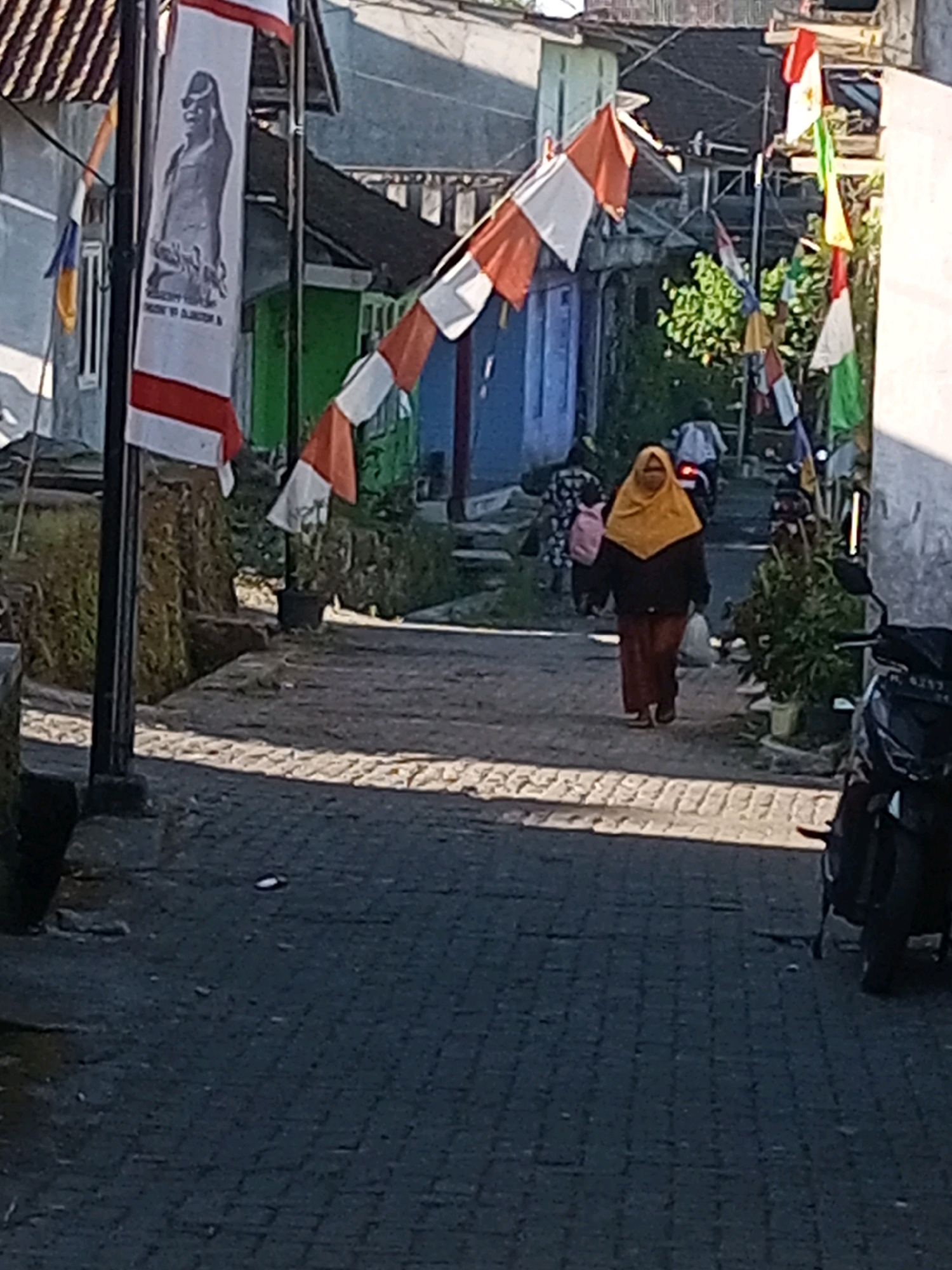 The image depicts a narrow, paved alleyway in Andongsili, Indonesia, lined with modest, colorful houses. A woman wearing a yellow hijab and brown pants walks away from the camera down the alley. She is partially obscured by the shadow of a nearby building. Several flags are visible, including a large banner with a black and white image of a figure and red and white stripes, as well as smaller red and white checkered flags strung across the alley. A motorcycle is parked on the right side of the alley, its black body and silver accents clearly visible. Another person, possibly a child carrying a pink backpack, is walking in the distance ahead of the woman in yellow. The scene appears to be outdoors, with sunlight casting shadows on the paved ground, suggesting it is daytime. The buildings exhibit simple architectural styles with visible pipes and wires.