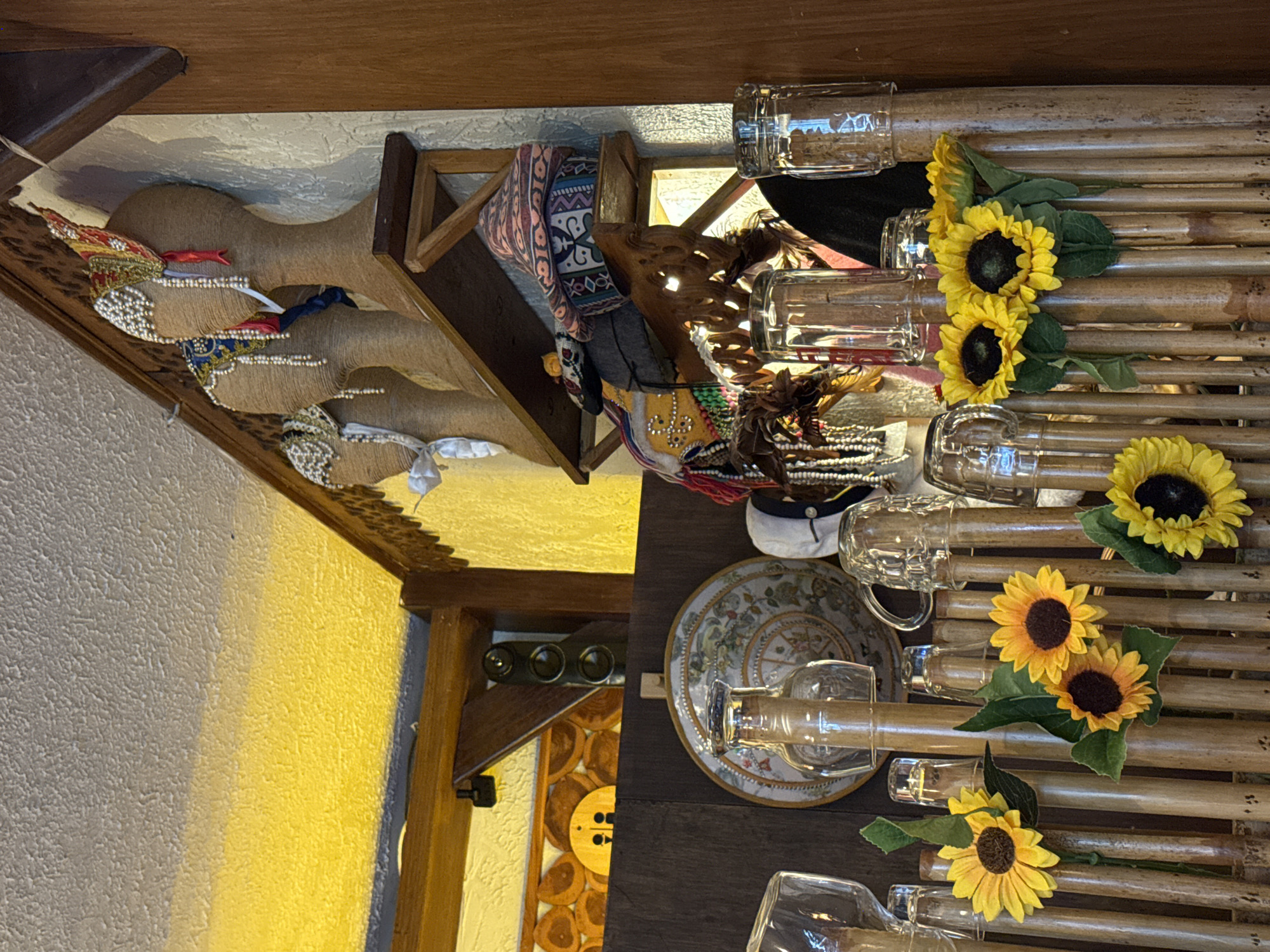The image shows a room in Baan Chaweng Noi, Thailand. A variety of decorative objects are arranged on shelves or displayed on a table. These include handcrafted dolls or figures adorned with headdresses and embellishments, a decorated plate, decorative objects including sunflowers, glass bottles placed on a bamboo-like surface, and various textiles and wooden elements. The room has a rustic or artisan-style aesthetic with a mix of textures and colors. The wall color varies from white to yellow.