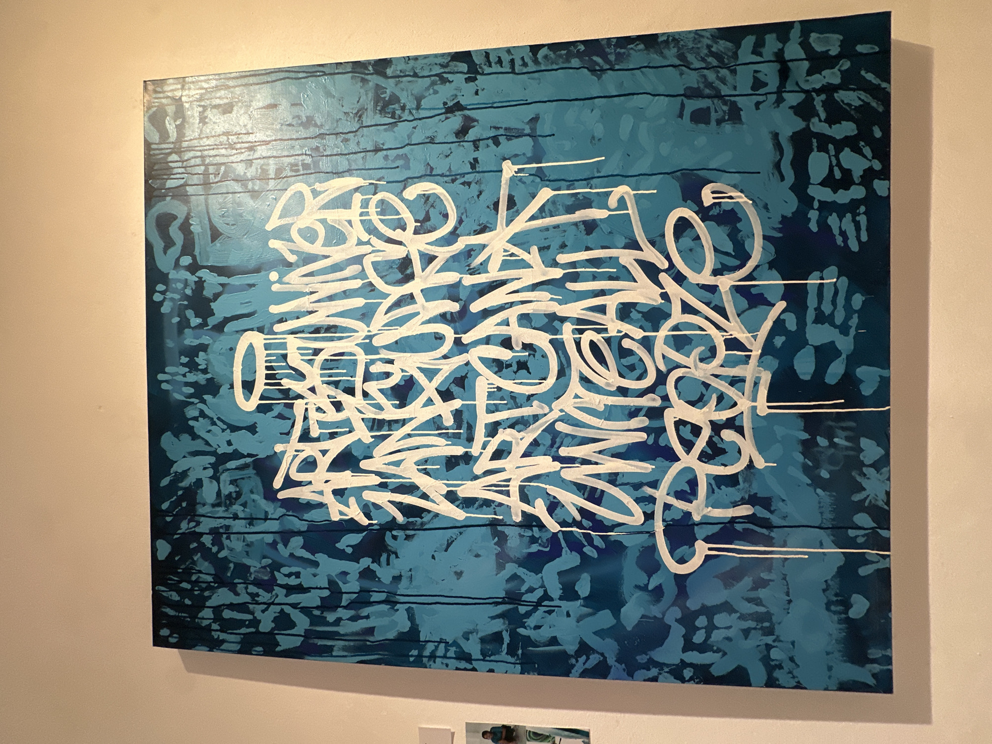 A close-up shot captures a large, vibrant abstract painting with graffiti-style lettering on a blue background. The painting features white, drippy lettering that appears to spell out "ART IS UNIVERSAL LANGUAGE ART CAN UNITE ALL PEOPLE". The background is a textured blend of dark and light blues, with vertical streaks that resemble dripping paint. The artwork is mounted on a light-colored wall, with a sliver of another framed piece of art visible to the left. The lighting in the room is warm and casts a shadow on the wall below the painting. The scene appears to be indoors, possibly in a gallery or home.