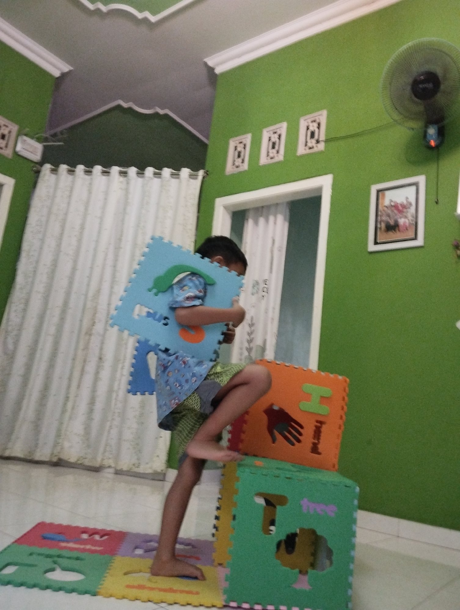 A young child is playing with foam alphabet puzzle pieces in a room with green walls, possibly in Pakembaran, Indonesia. The child is standing on a yellow puzzle piece and holding a blue puzzle piece that depicts a green caterpillar. The child is wearing a blue shirt with a pattern and green shorts. In the background, white curtains hang from a window, and a ceiling fan is mounted on the wall. Other puzzle pieces, including an orange one with the letter 'H' and a hand, and a green one with the letter 'T' and a tree, are stacked on the floor. The floor is tiled with a light-colored pattern. The room appears to be a play area or a child's bedroom.