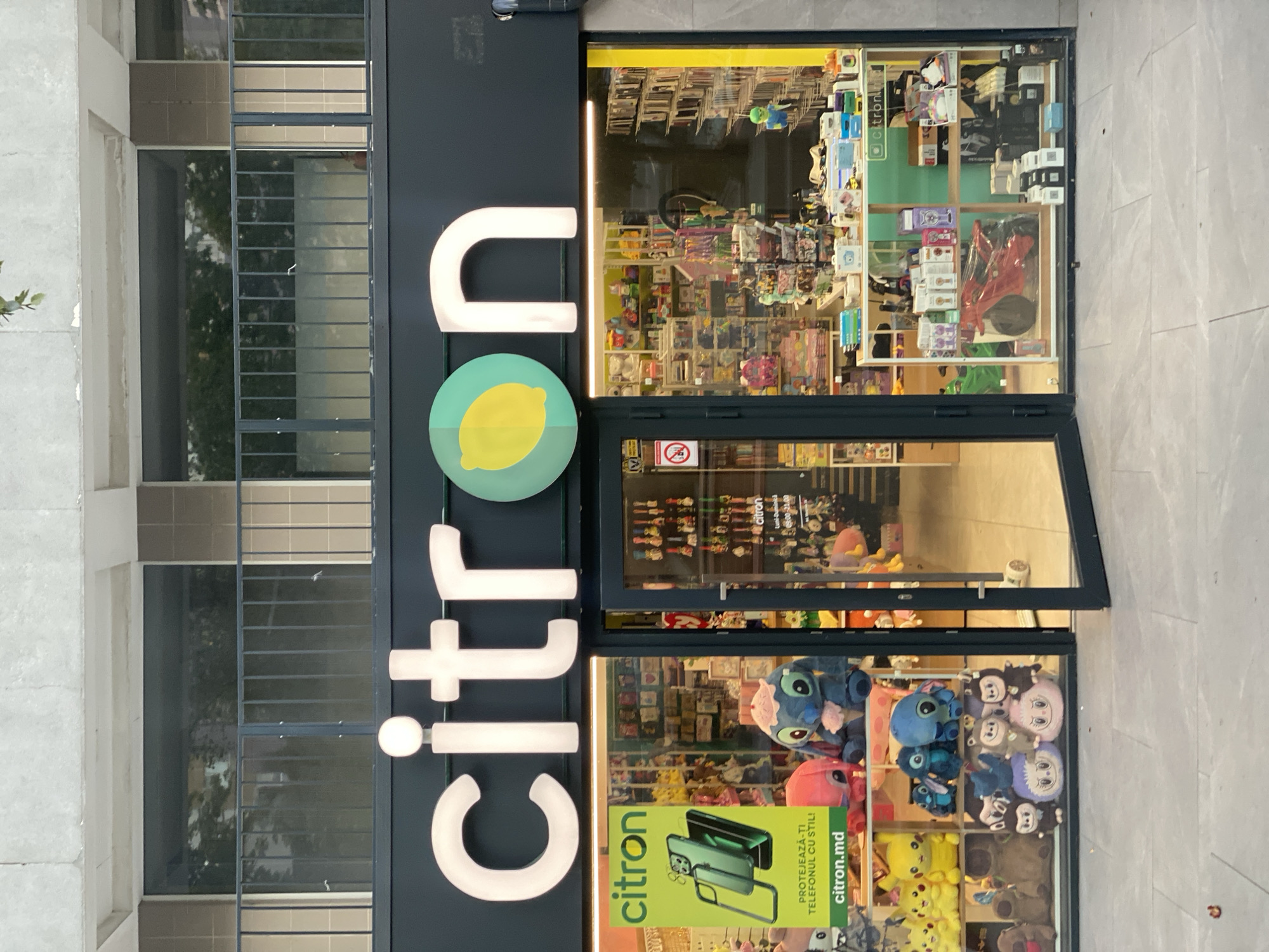The image shows the storefront of a "Citron" store located in Chișinău, Moldova. The store has a large illuminated sign with the word "citron" and a lemon graphic. The storefront window displays a variety of merchandise, including plush toys, phone accessories, and other retail items. Inside the store, shelves are filled with products, and a red toy motorcycle is visible. The entrance to the store is open, and a closed glass door is to the right of the entrance. The exterior of the building above the storefront features a concrete facade with windows and a railing. The ground in front of the store is paved with large gray tiles.