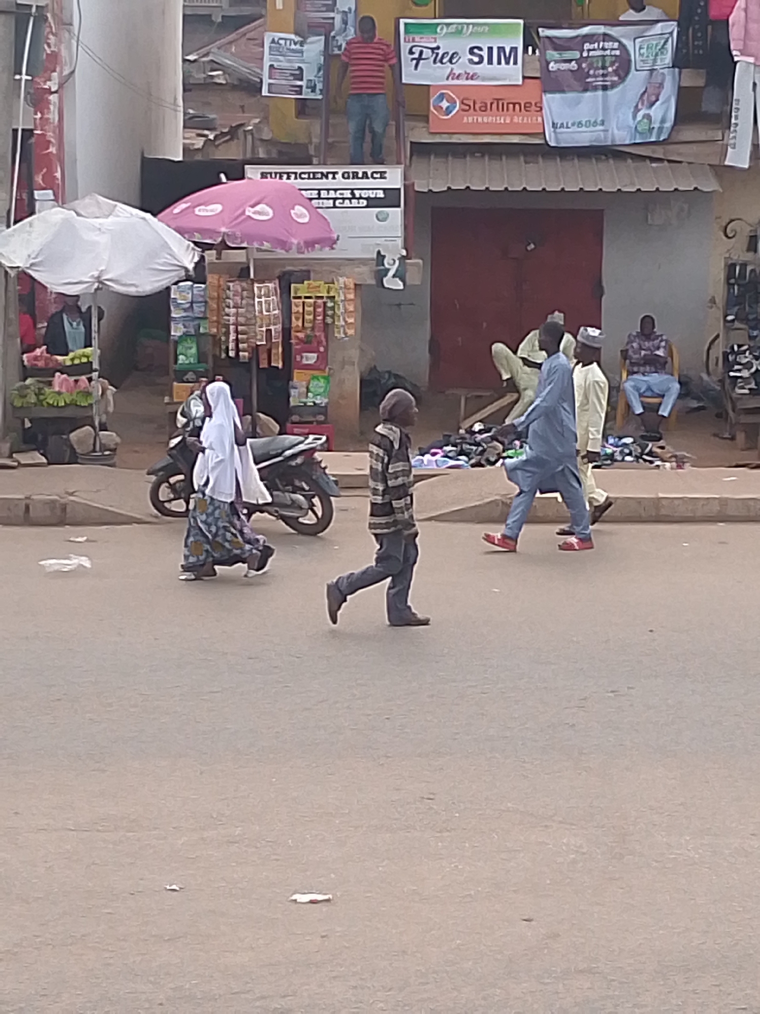 The scene shows a street in Bukuru, Nigeria. Pedestrians, including a woman in a white hijab, a man in a striped shirt, and two men in traditional robes, are crossing the street. A motorcycle is parked near a stall selling goods under pink and white umbrellas. Several men are seated near a building displaying signs for "Free SIM," "StarTimes," and "SUFFICIENT GRACE." A man stands on a ladder next to these signs. A fruit stand is visible on the left side of the image. The street appears to be paved and fairly empty of vehicles.