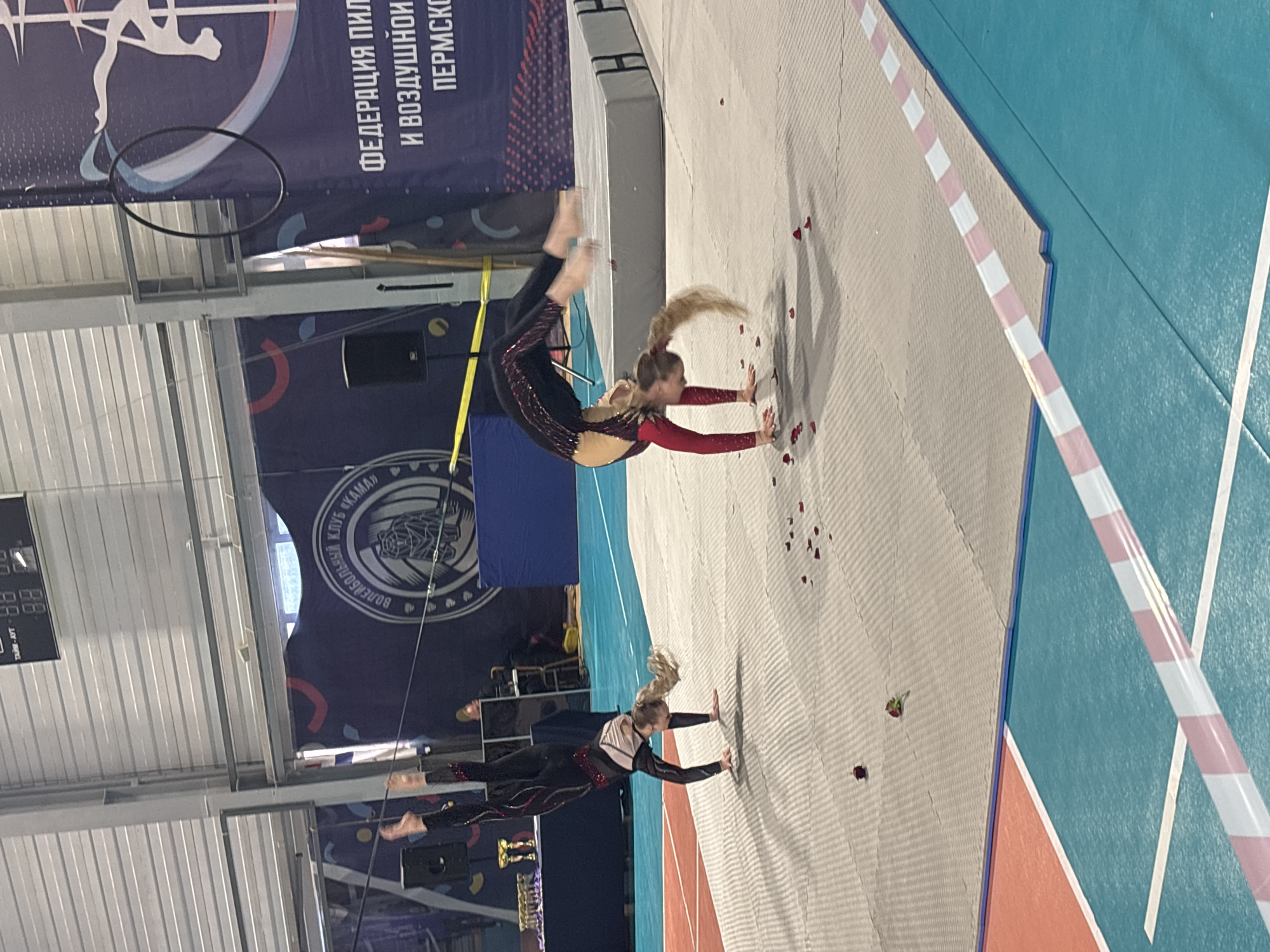 In an indoor sports arena in Perm, Russia, two young female gymnasts perform handstand variations on a light-colored padded mat. The gymnast on the left, wearing a black and red leotard, is executing an upright handstand. The gymnast on the right, in a red and black leotard, is performing a handstand with an arched back and bent legs. Red rose petals are scattered across the mats around them.
In the background, to the left, a dark blue table holds several gold and silver trophies. A black speaker on a stand is positioned nearby. Centered in the background is a large dark blue banner displaying "ВОЛЕЙБОЛЬНЫЙ КЛУБ «КАМАЙ»" (Volleyball Club "Kamay") with a stylized animal logo. To the right, another large dark blue banner partially reads "ФЕДЕРАЦИЯ ПИЛ И ВОЗДУШНОЙ ПЕРМСКО" (Federation of Pole and Aerial Perm), with a black aerial hoop hanging in front of it. A grey crash mat is positioned behind the right gymnast.
The floor of the arena features sections of blue court with white lines and a reddish-orange area. A pink and white striped tape marks a boundary line on the blue court. Overhead, a scoreboard displays "ТАЙМ - АУТ" (TIMEOUT) and numbers "88 88". The ceiling consists of corrugated metal panels. Shadows are visible beneath the gymnasts, cast by overhead lighting. A partial view of a person wearing a yellow hat is visible behind the background table.