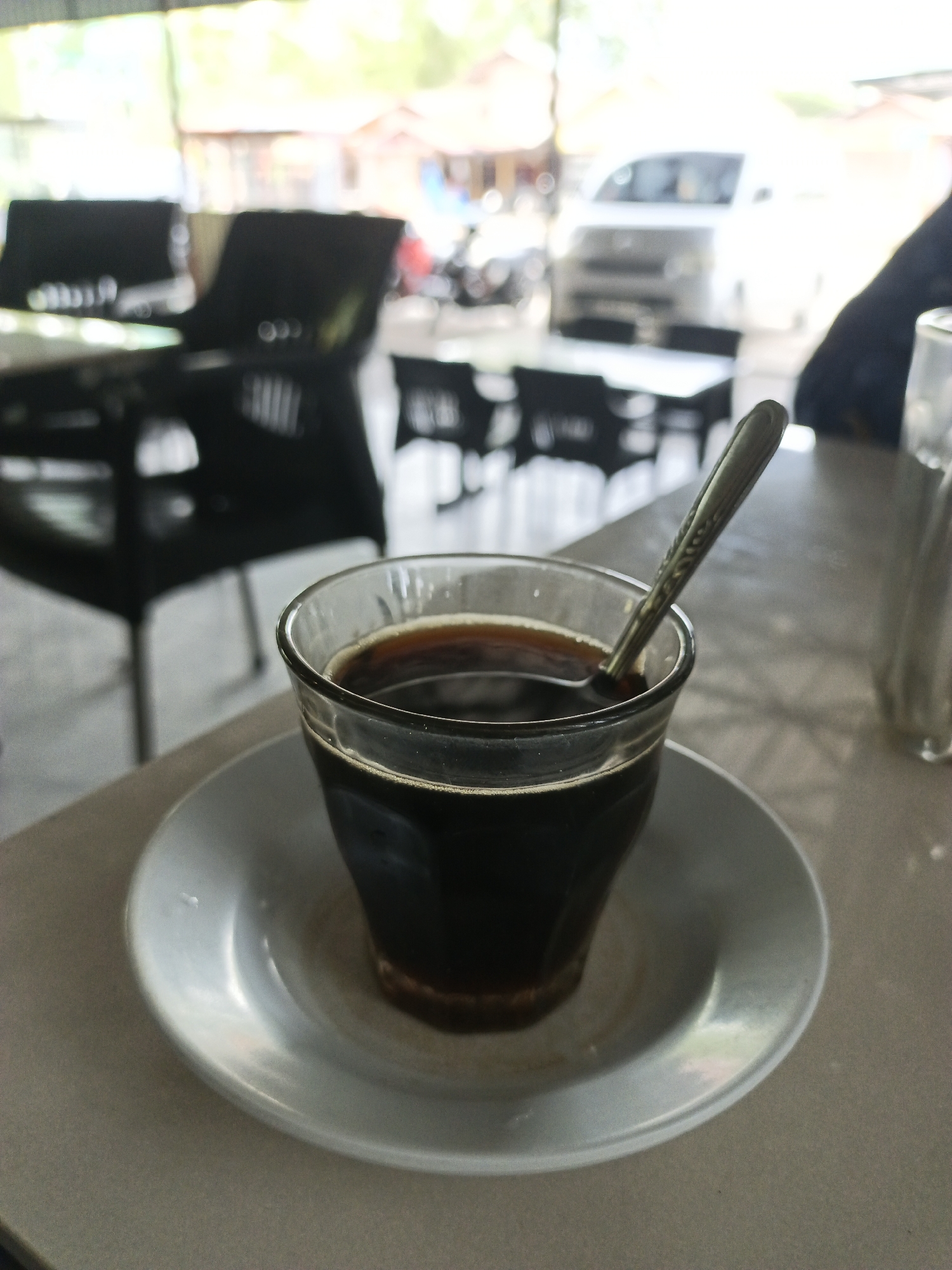 A clear glass of dark coffee sits on a white saucer on a tan-colored table. A small spoon is inside the glass. In the blurry background, black chairs and tables are visible. Outside the building, there is a white vehicle and a motorcycle. The location is Lhokseumawe, Indonesia.