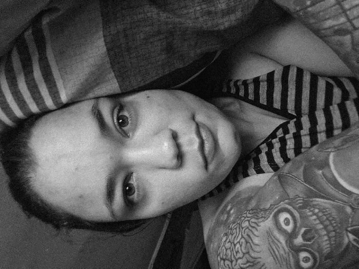 This black and white photograph depicts an individual, appearing to be an adult female, in a close-up, tilted shot. Her head is positioned centrally, resting on a striped pillow or blanket featuring alternating dark and light bands. She is wearing a garment with a similar striped pattern on her upper body. Her eyes are open, directed towards the camera with a neutral expression. To the left and behind her head, another pillow or bedding item is visible, adorned with a prominent graphic of a stylized skull with large, wide-open eyes and brain matter exposed on its crown. The background is dark and undefined, indicating an indoor setting, likely a domestic bedroom. The image provides no specific visual information to determine a precise location within Buenos Aires, Argentina.
