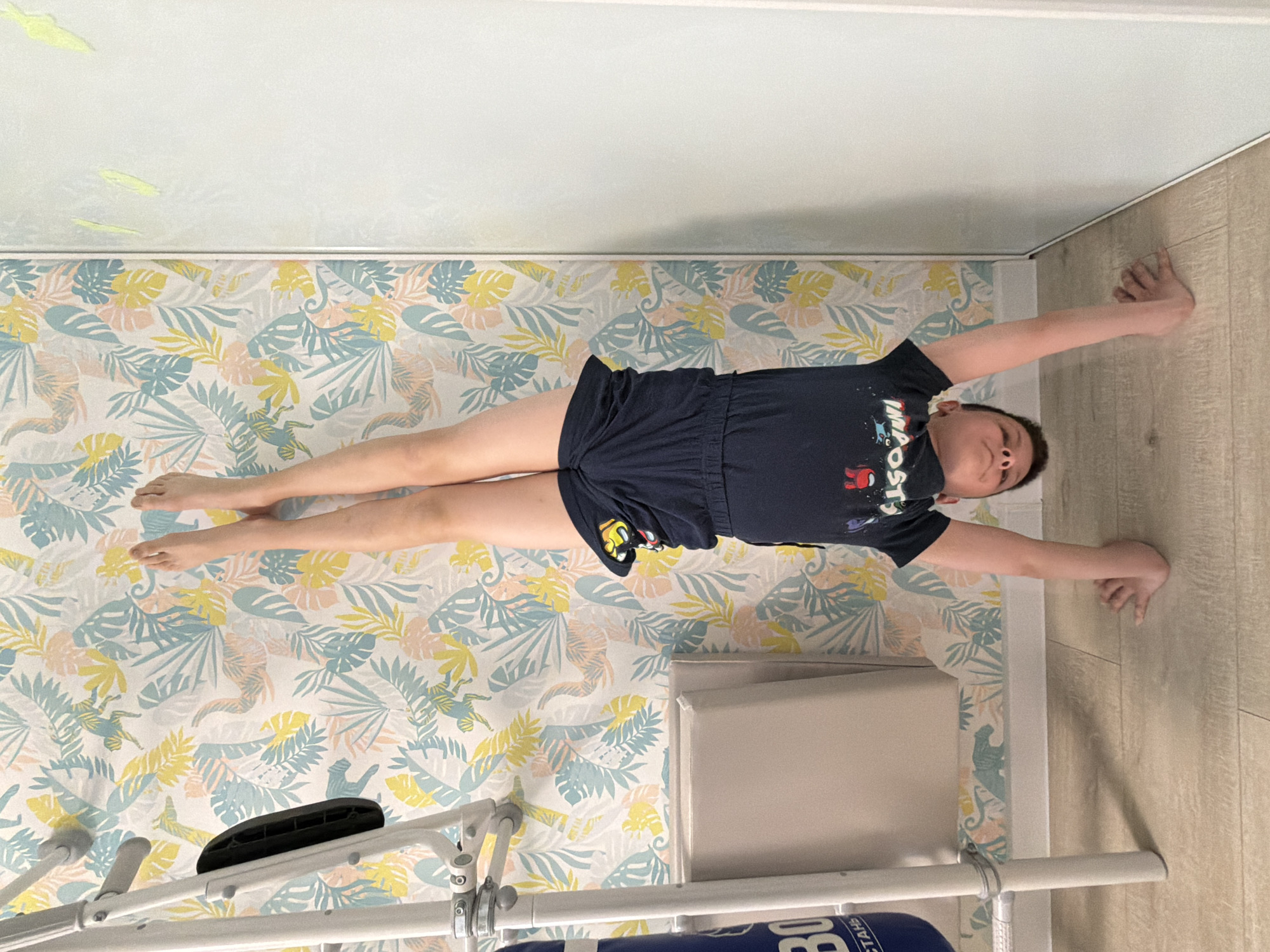 A child lies supine on a patterned surface in what appears to be a room in Votkinsk, Russia. Their arms are extended, hands touching a wall. The surface beneath them is decorated with a tropical leaf pattern in pastel colors. Adjacent to the patterned surface is a beige-colored floor. A piece of exercise equipment is visible at the bottom of the image. The walls of the room are a pale color.