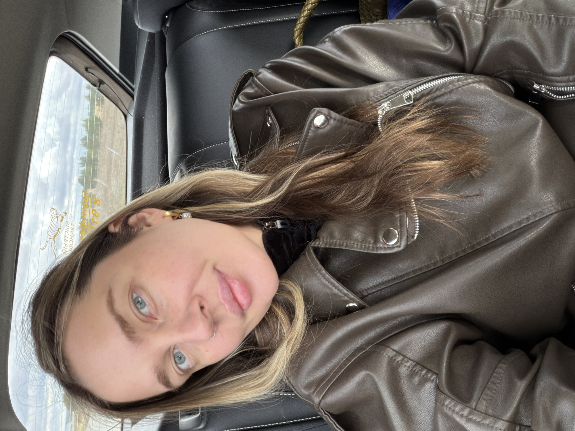 A woman is in the back seat of a car, wearing a brown leather jacket. The scene outside the window shows a field with some trees and buildings in the distance. In the window there is some text in a foreign language which is likely Russian. Based on that information, the location is likely in Povarenki, Russia.