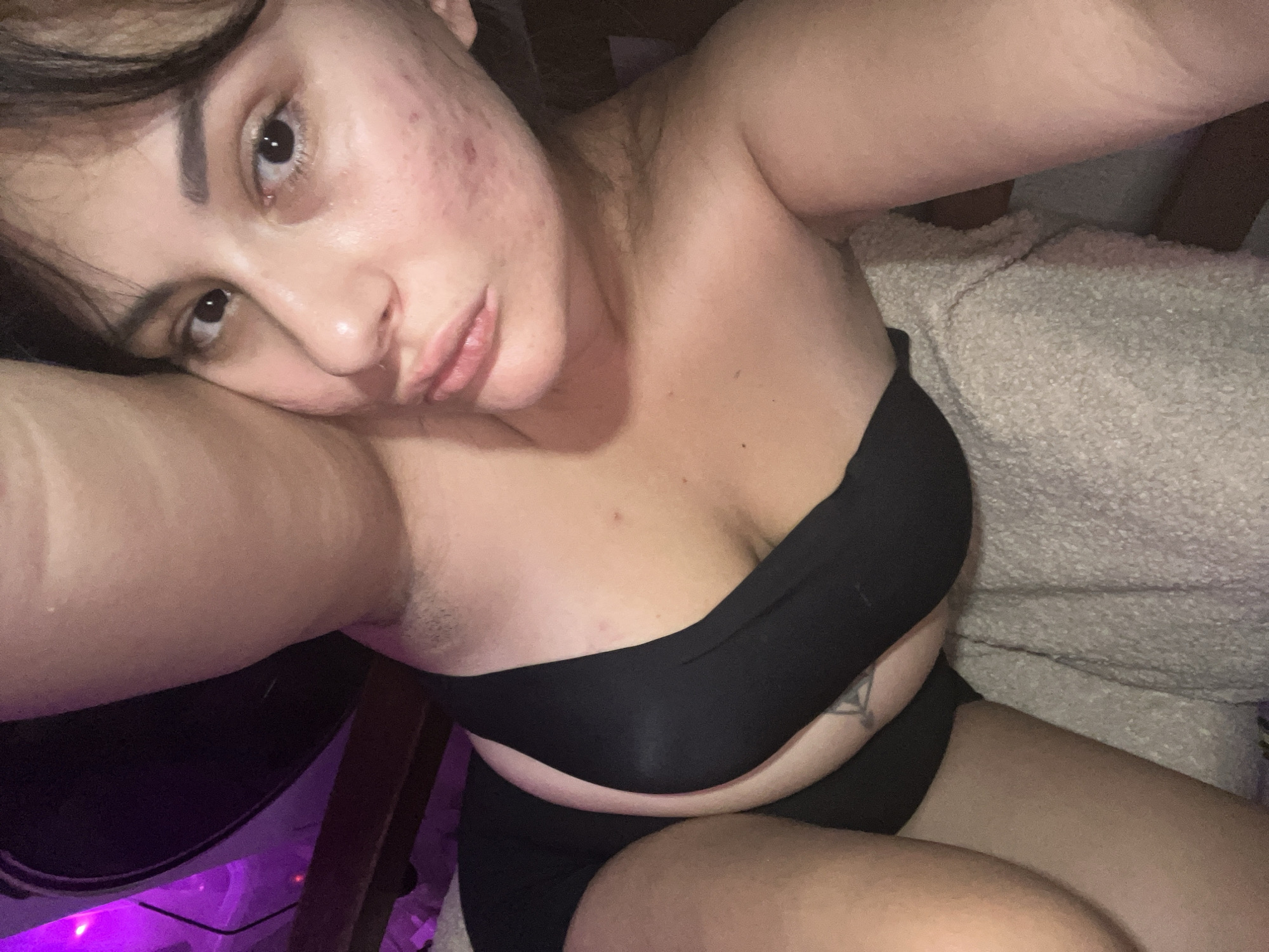 The image shows a close-up, selfie-style shot of a young woman lying down. She is wearing a black bra and black shorts. The lighting is dim and appears to be from a purple-colored light source in the background, possibly an LED strip or ambient lighting. The woman's face is partially visible, with her eyes open and looking towards the camera. Her skin has visible blemishes. Her arm is raised and bent, obscuring part of her body. She is lying on a textured blanket. There are no discernible elements in the image that would allow for the identification of a specific location in Kiryat Bialik, Israel.