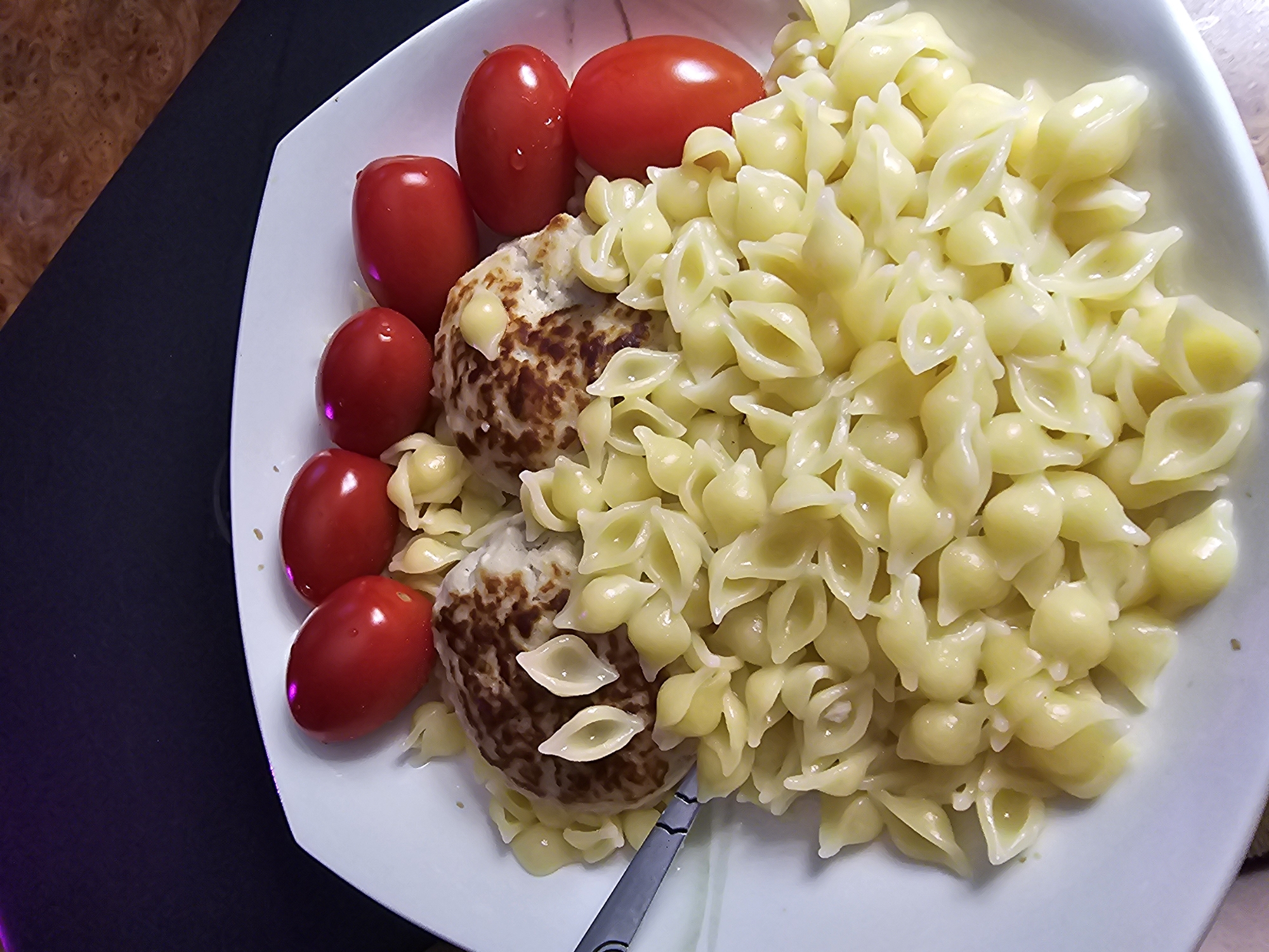 A close-up, overhead shot features a white plate of food on a dark surface. The plate contains a portion of shell pasta, two cooked hamburger patties, and several small, red cherry tomatoes. The pasta is a pale yellow and appears to be coated in a light sauce. The hamburger patties are round and speckled with brown, suggesting they are well-cooked. The cherry tomatoes are clustered to the left of the plate, with some showing a slight sheen that may be water droplets. A fork with a metallic handle is visible at the bottom right, partially submerged in the pasta. The background is out of focus, showing a dark, possibly black tablecloth, with a hint of a wood-grain pattern and purple lighting on the left side. The scene appears to be an indoor setting, likely a meal. No text is visible.
