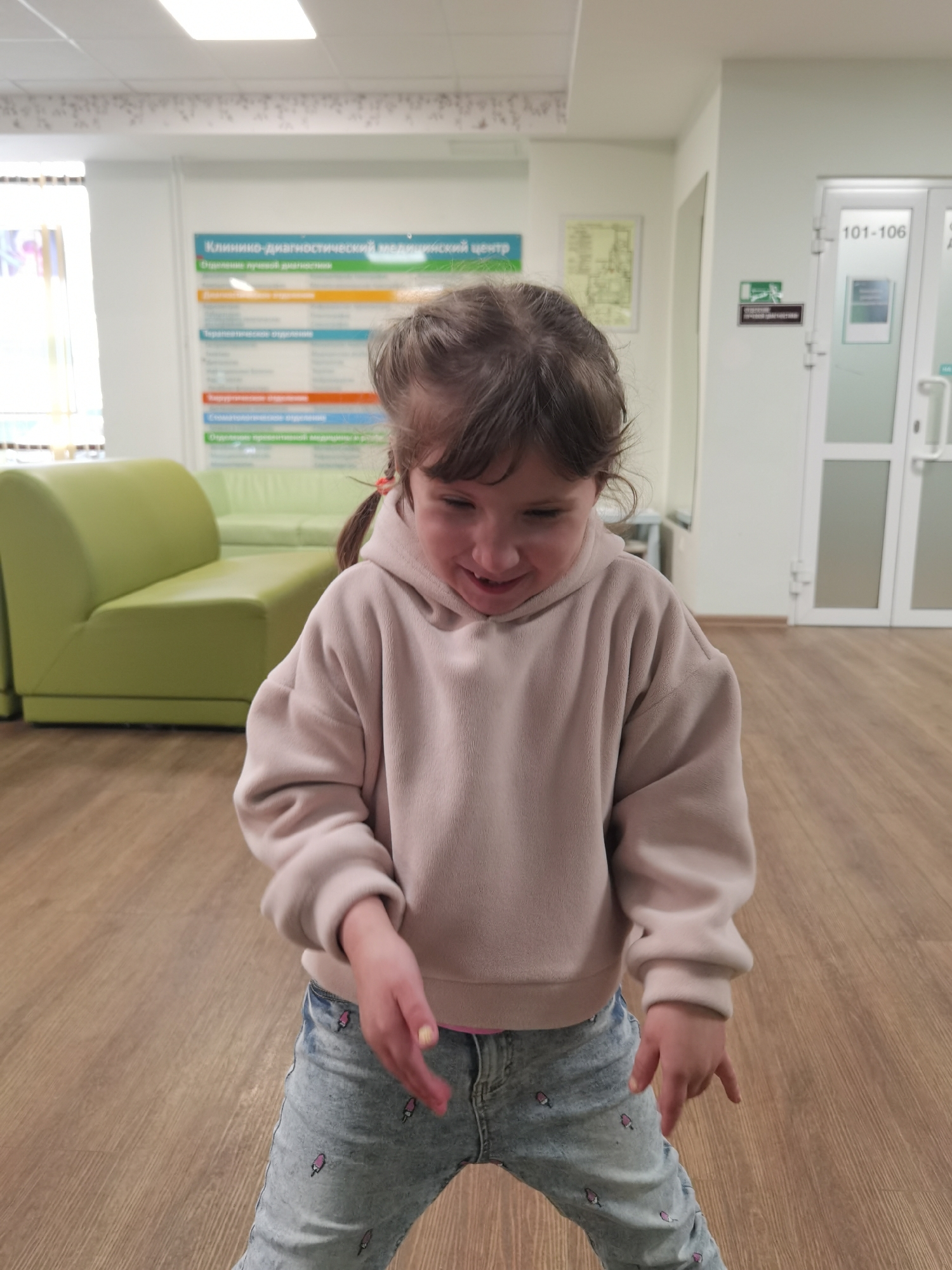 The image depicts a young female child, approximately 4-7 years old, standing in what is identified as a medical facility waiting area in Izhevsk, Russia. The child has dark brown hair styled in two pigtails secured with orange hair ties. She is wearing an oversized light beige fleece hoodie and light blue denim jeans patterned with small pink ice cream cones. She looks downwards and slightly to her left, exhibiting a closed-mouth smile or giggle, with her hands held loosely at waist height, fingers slightly curled.
The environment features light brown wood-patterned flooring and off-white walls. A decorative border with a subtle white floral design runs along the upper wall section. In the left background, a bright green modular sofa provides seating, and a large window with vertical blinds is visible behind it. In the right background, a set of white double doors with frosted glass panels is marked with room numbers "101-106" and an emergency exit sign displaying a green running person pictogram and Cyrillic text.
A prominent sign on the back wall, to the right of the child, is written in Cyrillic and identifies the location as a "Клинико-диагностический медицинский центр" (Clinical-Diagnostic Medical Center). Below the main title, various departments are listed on colored horizontal bars, including "Отделение лучевой диагностики" (Department of Radiation Diagnostics), "Терапевтическое отделение" (Therapeutic Department), "Хирургическое отделение" (Surgical Department), "Стоматологическое отделение" (Dental Department), and "Отделение профилактической медицины и реабилитации" (Department of Preventive Medicine and Rehabilitation). A framed diagram or map is also present on the wall. The ceiling contains recessed rectangular light fixtures.