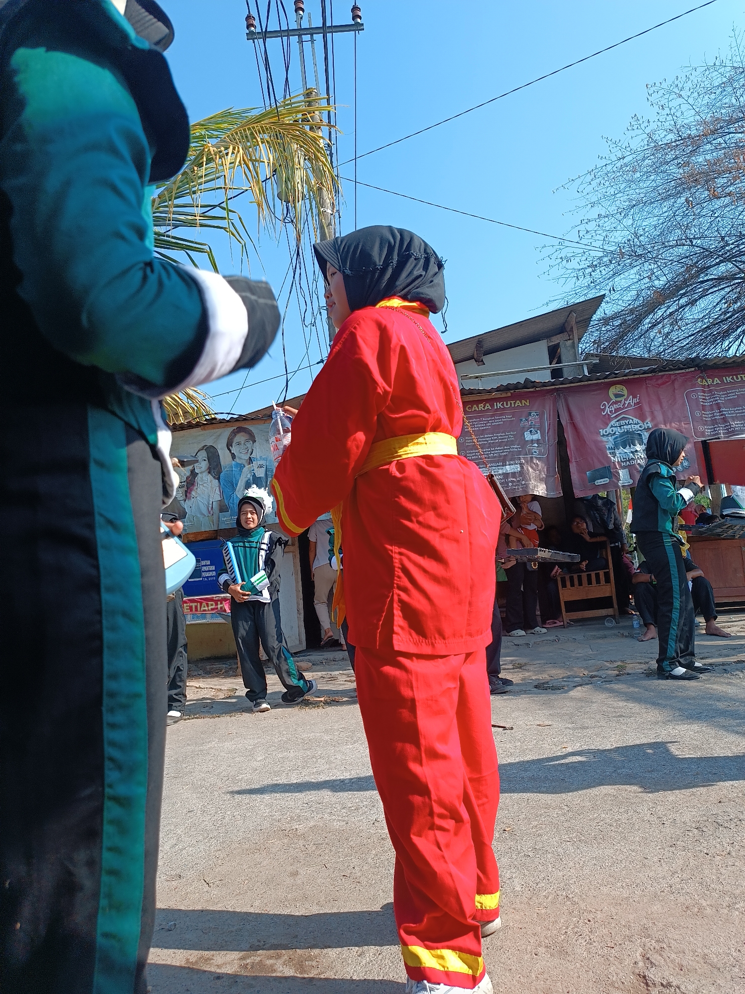 The image captures an outdoor scene, possibly in Imogiri, Indonesia, under a clear blue sky. In the foreground, a person wearing a teal and black uniform is partially visible from the back, their arm extended towards the right. The main subject in the mid-ground is a person dressed in a bright red uniform with a yellow sash, also seen from the side. This individual is wearing a black hijab. Behind them, another person in a teal and black uniform, presumably a child, stands with hands clasped, looking towards the right. Further back, a banner with Indonesian text and images is displayed. The background shows buildings and trees, with utility poles and wires overhead. The ground is paved with concrete or asphalt. The scene appears to be part of a public event or parade, given the uniforms and the banner.
