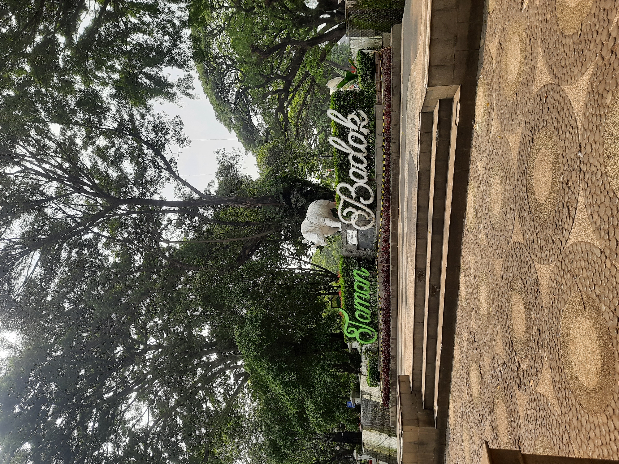 The image shows the entrance sign for Taman Badak in Bandung City, Indonesia. The sign consists of large, stylized green letters spelling "Taman" on the left and white cursive letters spelling "Badak" on the right. Behind the sign, a white rhinoceros statue is visible. The entrance is set in a park-like environment with lush green trees and landscaping. A paved walkway with circular stone patterns leads up to a set of steps. The sky is overcast.