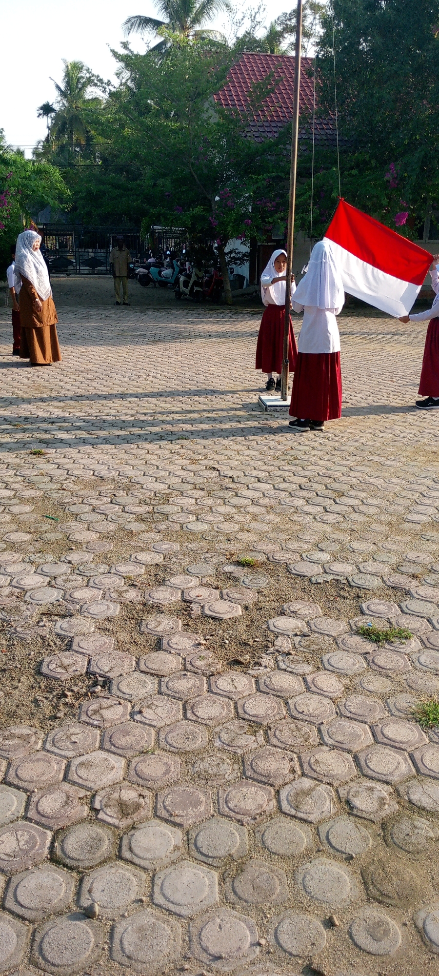 The image depicts an outdoor scene in Baratpayaitek, Indonesia, likely during daylight hours. In the foreground, a patterned pavement made of hexagonal concrete blocks covers the ground. Several individuals, appearing to be students wearing school uniforms consisting of white tops and maroon skirts or hijabs, are positioned around a flagpole. One student is actively raising a red and white Indonesian flag. In the background, lush green trees with some flowering plants are visible, along with a building with a red tiled roof. Further in the background, a man in a light-colored uniform stands near a gate, and several motorcycles are parked. The overall setting suggests a schoolyard or similar public space.