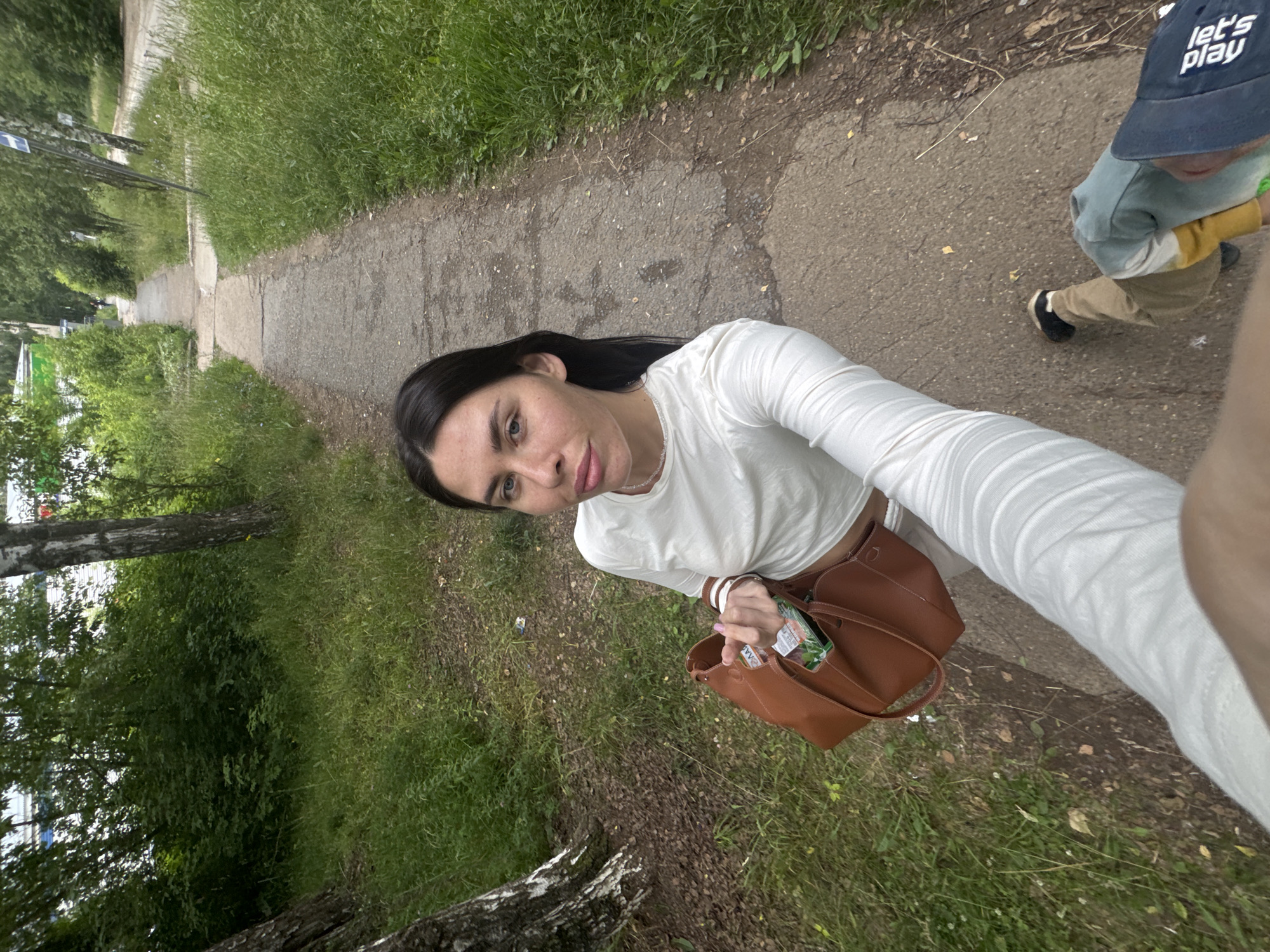 This image shows a young woman and a child walking along a paved path in an outdoor setting. The woman is in the foreground, taking a selfie. She has long, dark hair and is wearing a white, long-sleeved shirt and white pants. She is carrying a brown purse. The child is to her right, wearing a light blue shirt, tan pants, and a blue baseball cap that says "let's play." The path is surrounded by lush green grass and trees. The overall impression is of a casual outdoor stroll, possibly in a park or along a nature trail. The lighting suggests it is daytime, and the greenery indicates it is likely spring or summer. The text on the child's hat, "let's play," suggests a playful mood. The scene appears to be set in Chaikovsky, Russia, as per the provided location context.