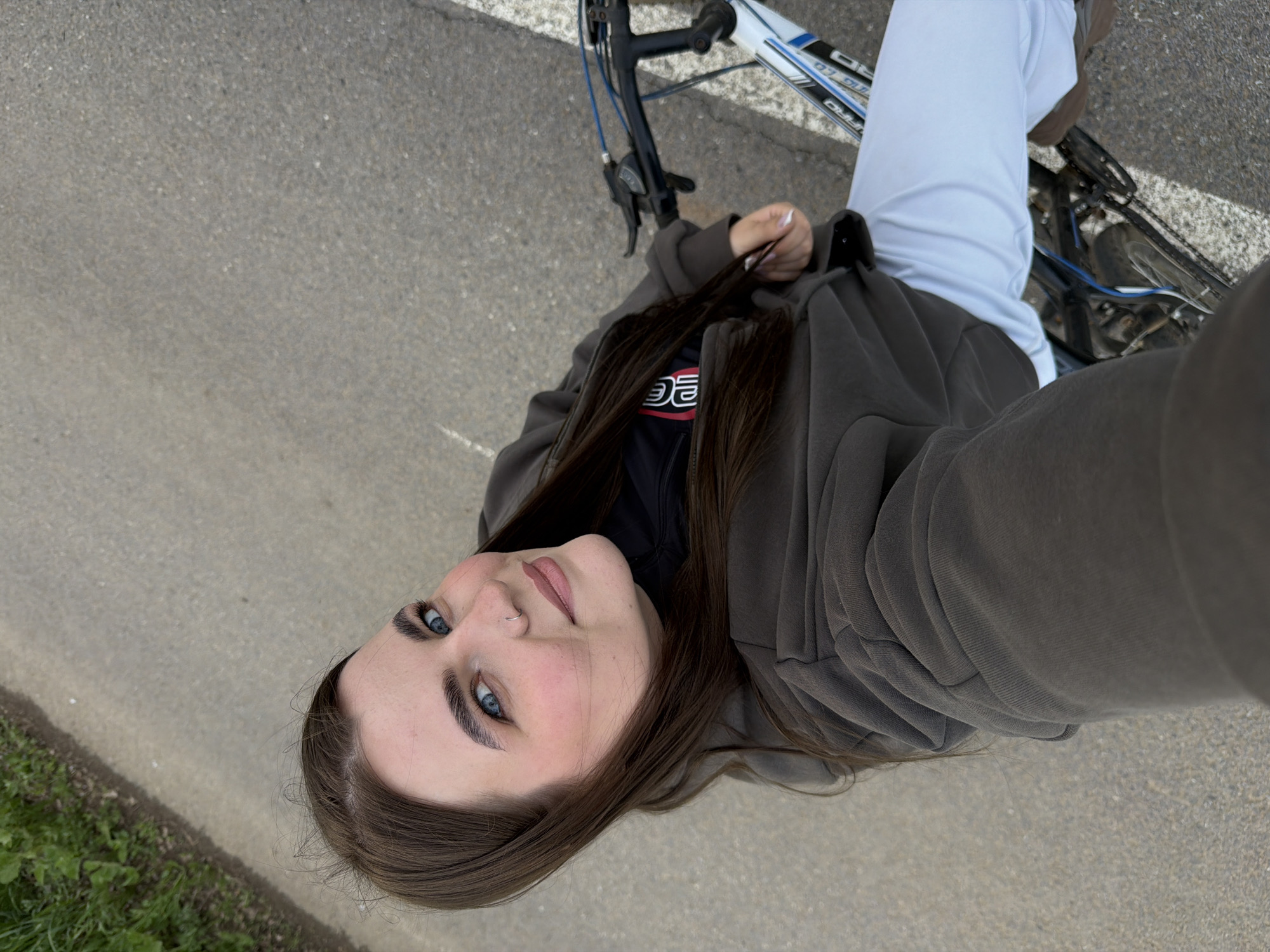 An overhead selfie captures a young woman outdoors in Marakushi, Russia, with her bicycle. She has long dark brown hair, bright blue eyes, and a small nose piercing, looking directly up at the camera with a calm expression. She is wearing a dark grey or brown oversized zippered hoodie over a black t-shirt, on which a red and white logo, possibly "DE" or "OG", is partially visible. Her light-colored pants are also visible.
Beside her is a bicycle with a white and blue frame; text on the frame includes "PRO" and other indiscernible letters. The scene is set on a grey, asphalt-like paved surface, with hints of sparse green grass and weeds along the edges of the path. The lighting suggests it is daytime, with soft, diffused light and no harsh shadows. The overall impression is casual and relaxed.