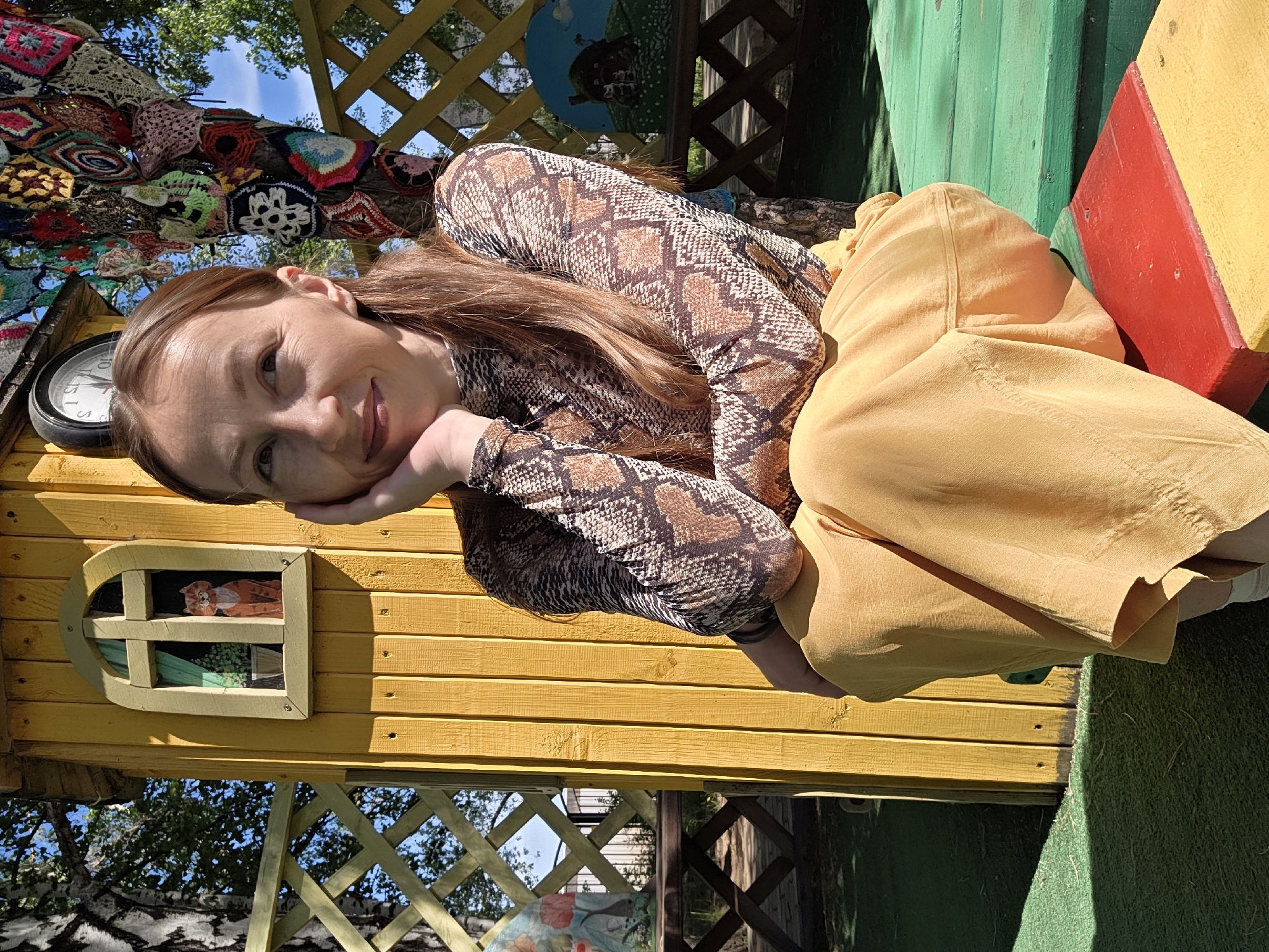 The image shows a woman with long brown hair wearing a snakeskin-print shirt and mustard-colored pants, resting her head on her hands and smiling. She is sitting in or against a brightly colored playhouse, painted in yellow, green, and red. The playhouse has a small window with a picture of a cat inside. Above the playhouse, there is a tree decorated with crocheted squares. The playhouse is located in the city of Chaikovsky, Russia. The overall setting suggests an outdoor environment, possibly a park or garden.