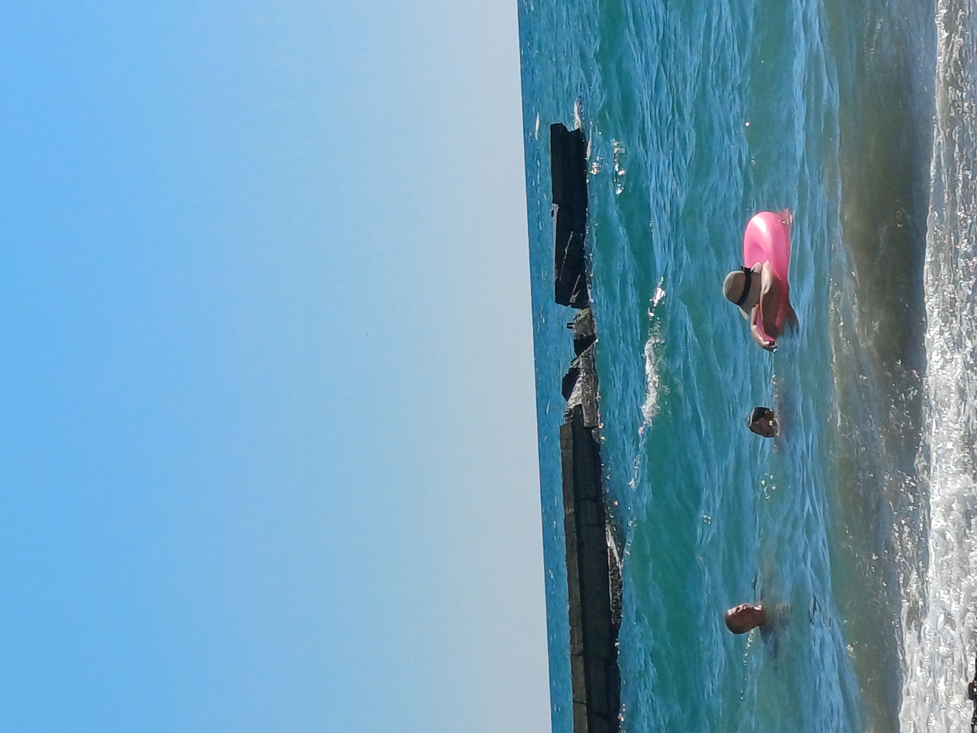 The image shows a sunny day at the sea, likely in Sochi, Russia, given the presence of a breakwater made of concrete blocks. The sea is a vibrant turquoise blue with moderate waves. In the foreground, the surf crashes onto a sandy or pebble beach.
There are three people visible in the water. On the left, a man with short, light-colored hair is swimming, with his head and shoulders above the water. He is looking towards the right. To his right and slightly further back, another person's head is visible above the water, appearing to be a child or a person with dark hair. In the mid-ground, a person wearing a wide-brimmed straw hat is floating on a bright pink inflatable ring. Their arms are resting on the ring, and they appear to be relaxed and enjoying the water.
The breakwater stretches across the middle ground of the image, extending from the left to the right side. It is a low, horizontal structure made of large, weathered concrete blocks, some of which appear to be broken or submerged, suggesting it is an older or damaged structure. The sky above is a clear, cloudless light blue. The overall scene depicts a leisurely day at the seaside.