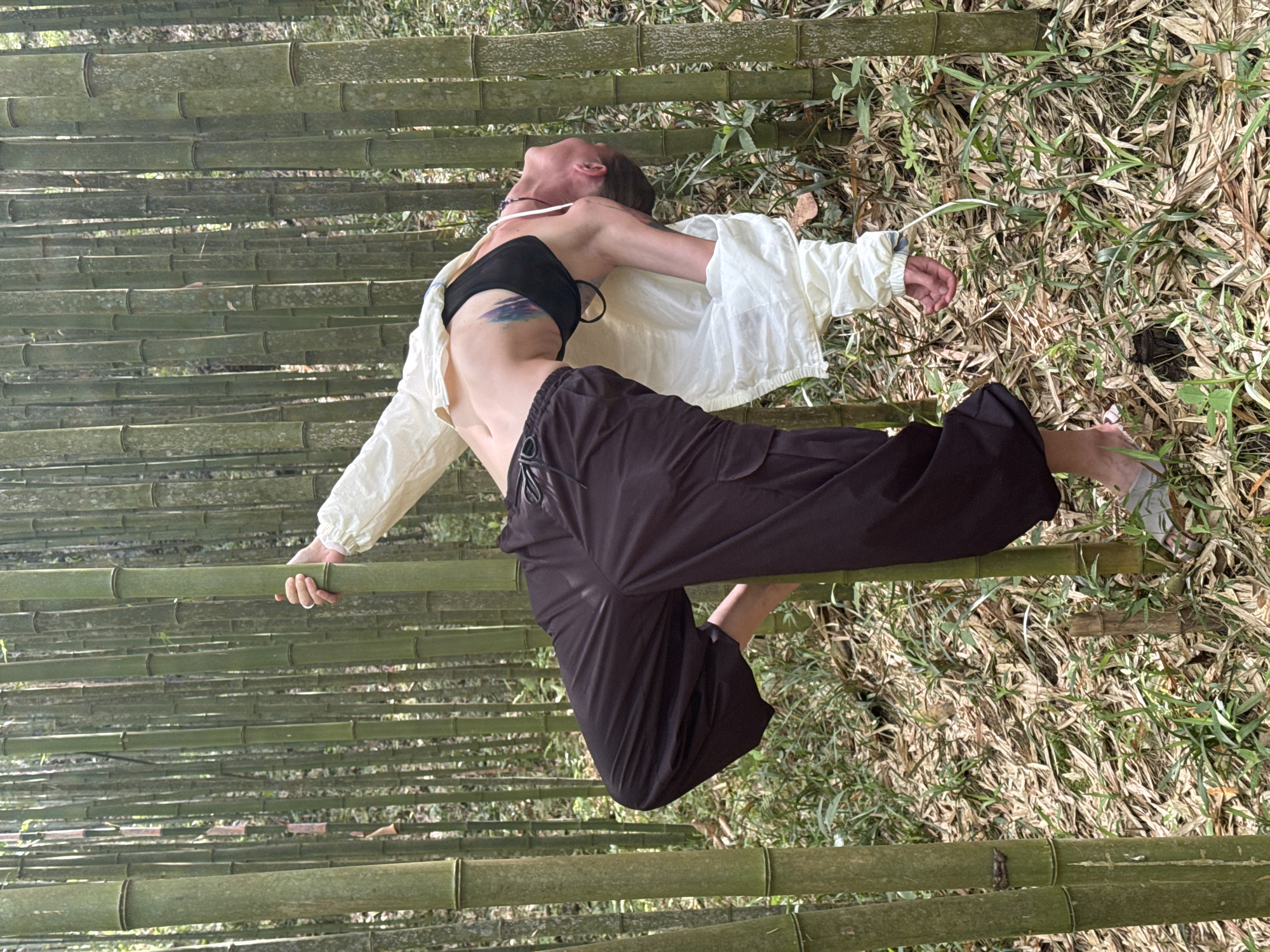 A woman is lying on a bamboo grove, her head tilted back. She wears a black strapless top, a light-colored open button-down shirt, and dark loose pants. The background is a mix of green bamboo stalks and dry vegetation on the ground. The location is suggested to be Ngã Ba Kim, Vietnam.
