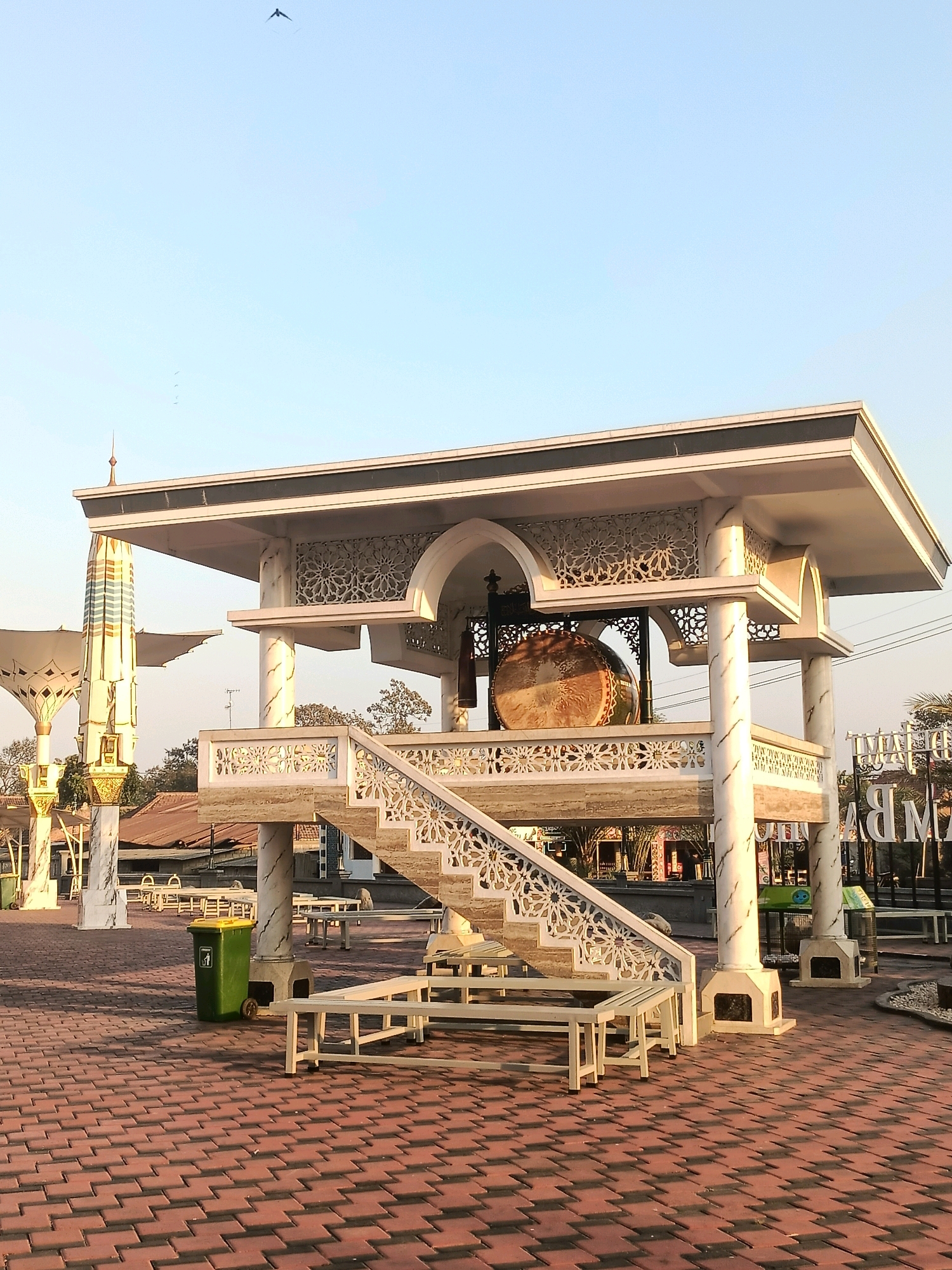 The image captures a sunny day in Plemahan, Indonesia, featuring an outdoor space with a prominent architectural structure. The structure, possibly a pavilion or a stage, is elevated and adorned with intricate white latticework. A large drum is visible within the structure, suggesting it might be a performance or ceremonial area. White benches are placed on the red brick paving in front of the structure, indicating a public gathering or relaxation spot. To the left, a tall, white umbrella-like structure with decorative elements stands, resembling a canopy for shade. In the background, a few trees and buildings with reddish-brown roofs are visible. The sky is a clear light blue with a few small birds in flight. A green trash bin is located on the paved area. The overall scene appears to be a well-maintained outdoor public space.