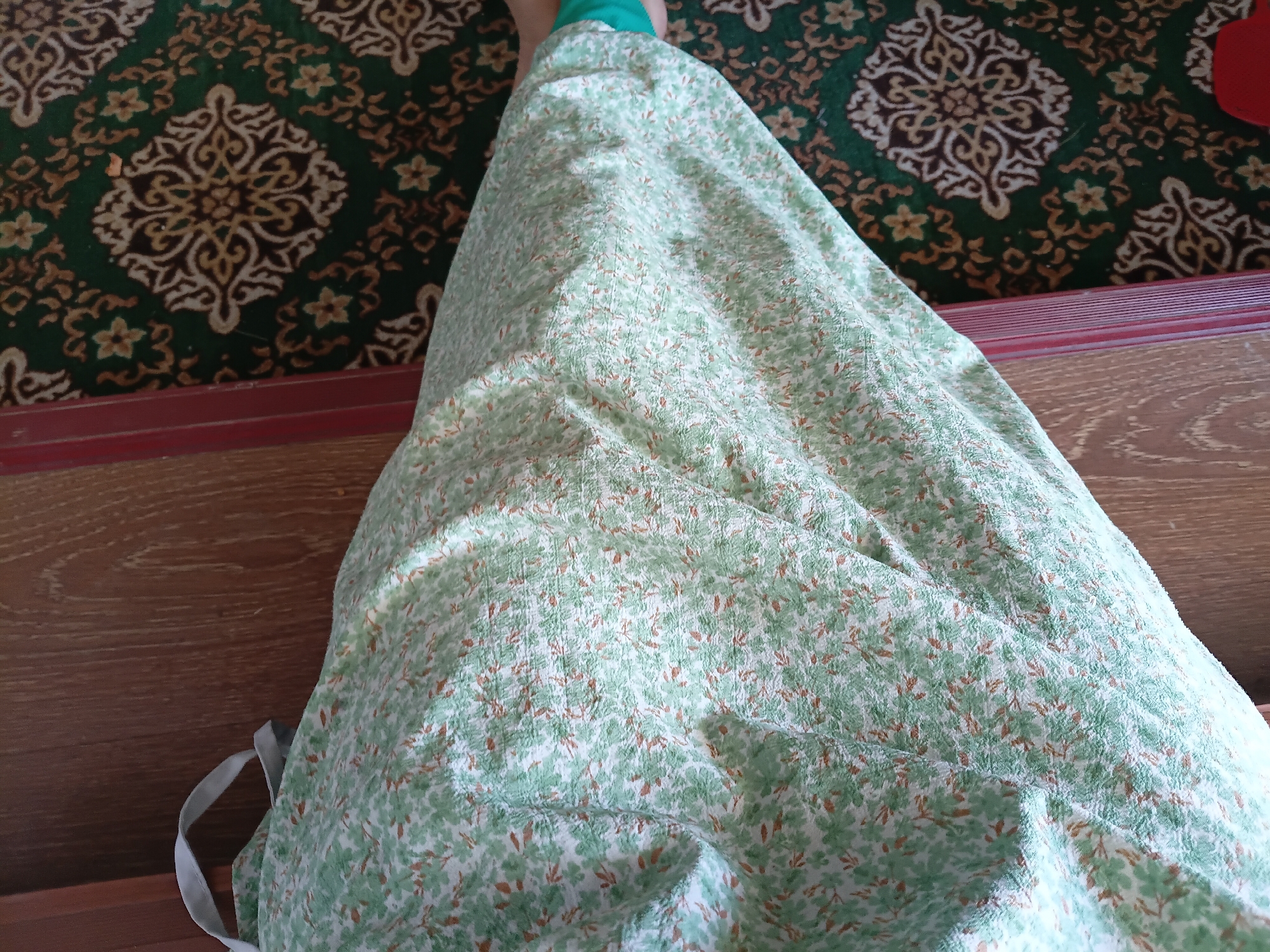 A close-up, top-down view of a person's legs covered by a light green, floral-patterned fabric, possibly a dress or skirt. The fabric is draped over the legs, creating folds and wrinkles. To the right of the fabric is a dark green carpet with an intricate, repeating pattern in beige and brown. A wooden surface, likely a floor or furniture, is visible to the left of the fabric. The scene appears to be indoors, possibly in a residential setting in Uchkun, Kyrgyzstan, given the context. There are no visible faces or identifying features of the person.
