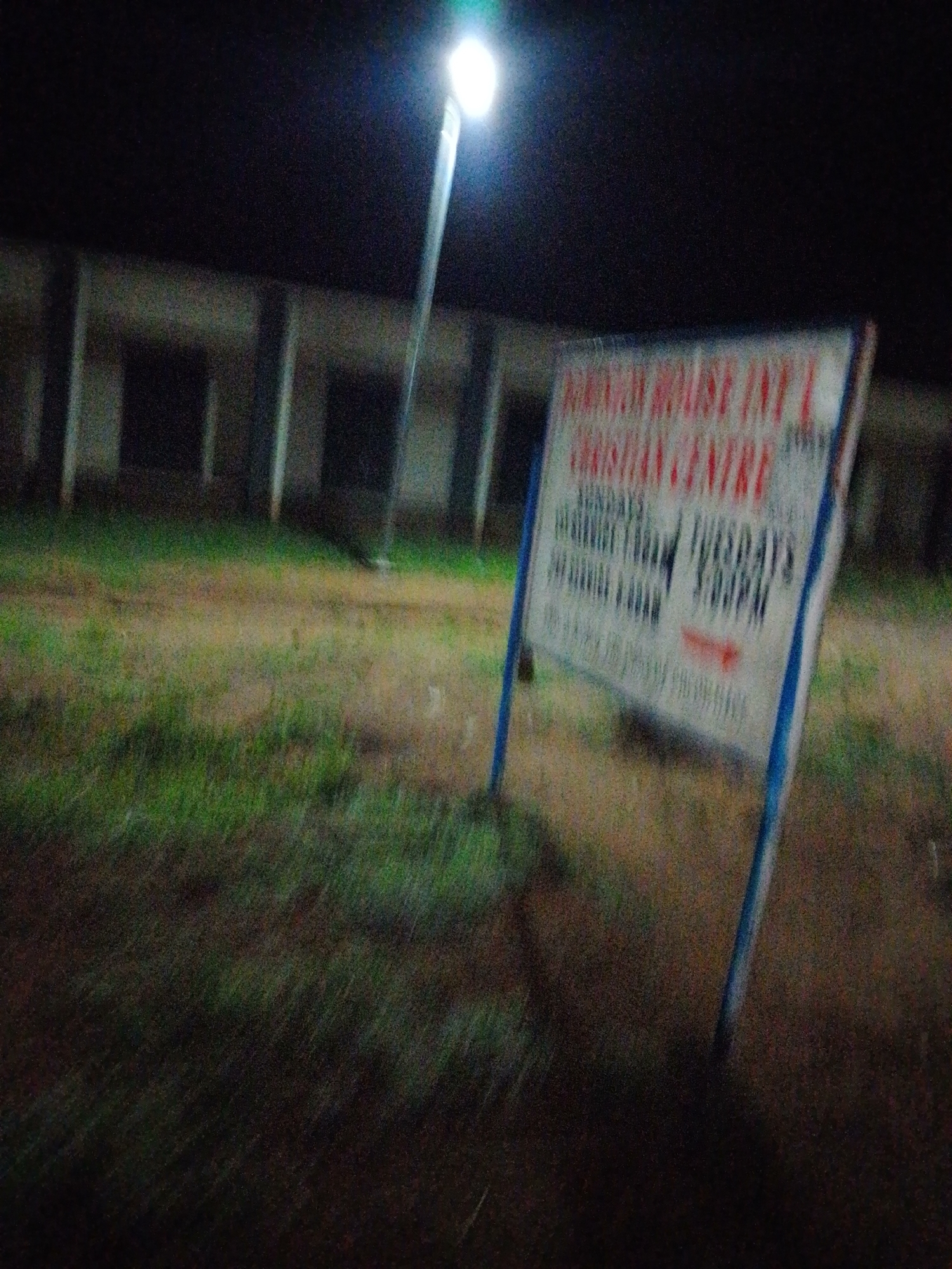 An outdoor, eye-level, low-light, motion-blurred photograph captures a signpost at night, likely in Jalingo, Nigeria, situated in front of a building structure with a grassy and dirt foreground. The signpost, predominantly white with red and blue lettering, advertises "KINGDOM HOUSE INT'L CHRISTIAN CENTRE." It lists service times, indicating "SUNDAYS: 8:00AM - 10:00AM" and "TUESDAYS: 5:00PM - 7:00PM." A street lamp illuminates the scene from the top left, casting a bright glow. The background is a dark, blurred representation of a building with visible columns. The overall image quality is degraded by graininess and motion blur. No individuals are clearly discernible in the frame.