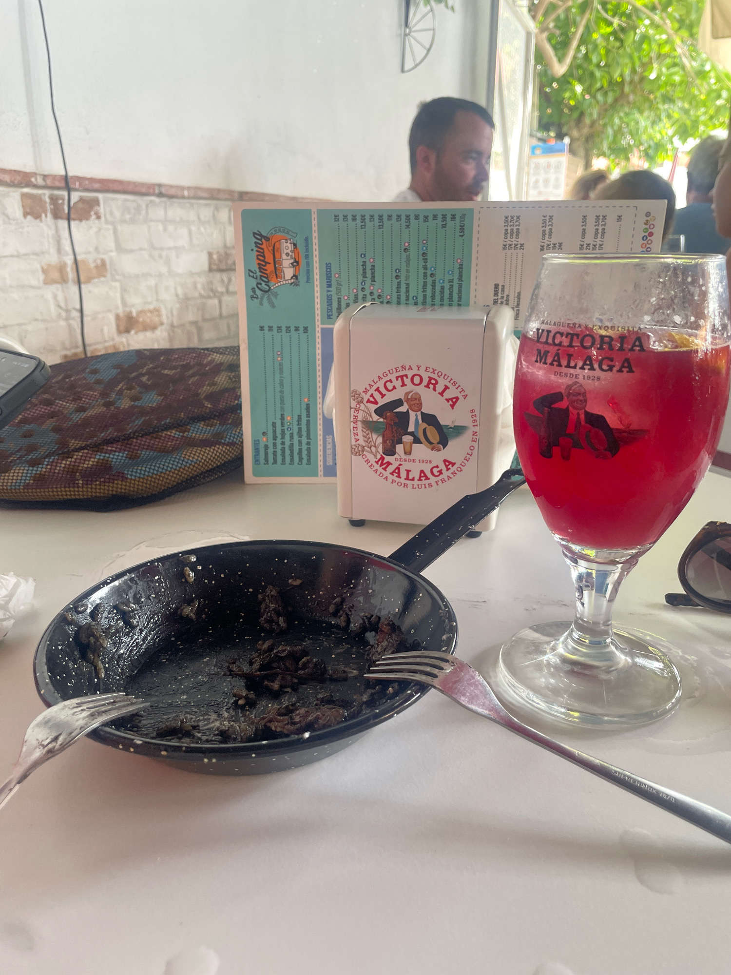 In La Herradura, Spain, a table is set for a meal. In the foreground, a black frying pan contains the remnants of a dark, saucy dish, with two forks resting nearby, one in the pan and another to the side. To the right, a tall stemmed glass holds a vibrant red beverage, likely a sangria, with a slice of orange visible. The glass is branded with "Victoria Malaga Desde 1928" and an image of a man in a suit. Adjacent to the glass is a white napkin dispenser, also bearing the "Victoria Malaga" logo. A menu, partially obscured, displays various food items and prices, suggesting a casual dining establishment. In the background, blurred figures are seated, with one man looking towards the camera. The setting appears to be outdoors or near an open space, with greenery visible and natural light illuminating the scene. The white walls and brick accents suggest a rustic or traditional architectural style.