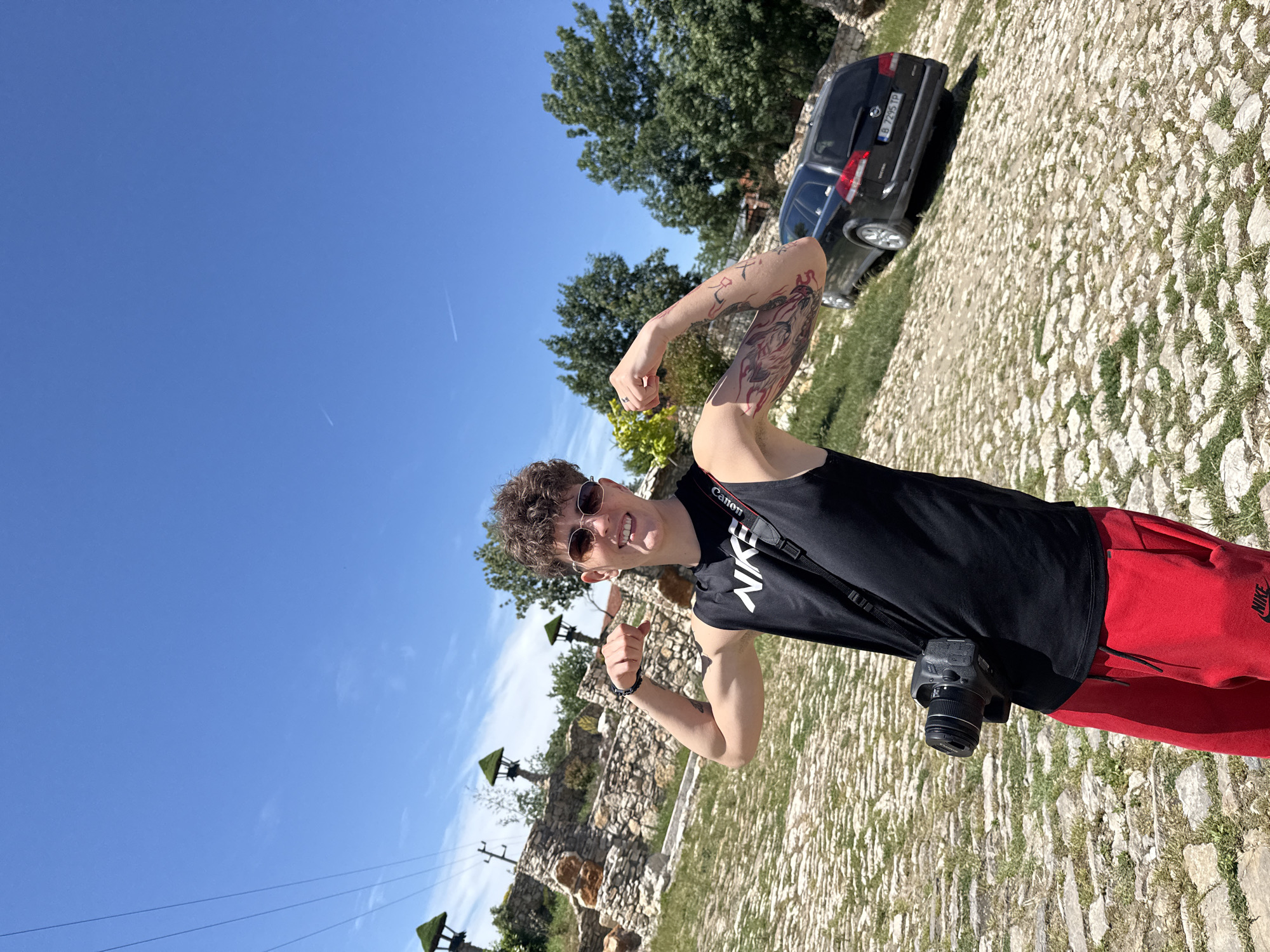 A young man stands on a cobblestone ground in the city of Lukovit, Bulgaria. He is wearing a black sleeveless shirt with white "NIKE" lettering, red sweatpants, and sunglasses. He has a Canon camera hanging around his neck. He is flexing his arms and smiling at the camera, giving two thumbs-up. His left arm is tattooed. Behind him is a black car with a Bulgarian license plate "B 1295 TP". There are green trees and a stone structure in the background under a clear blue sky.