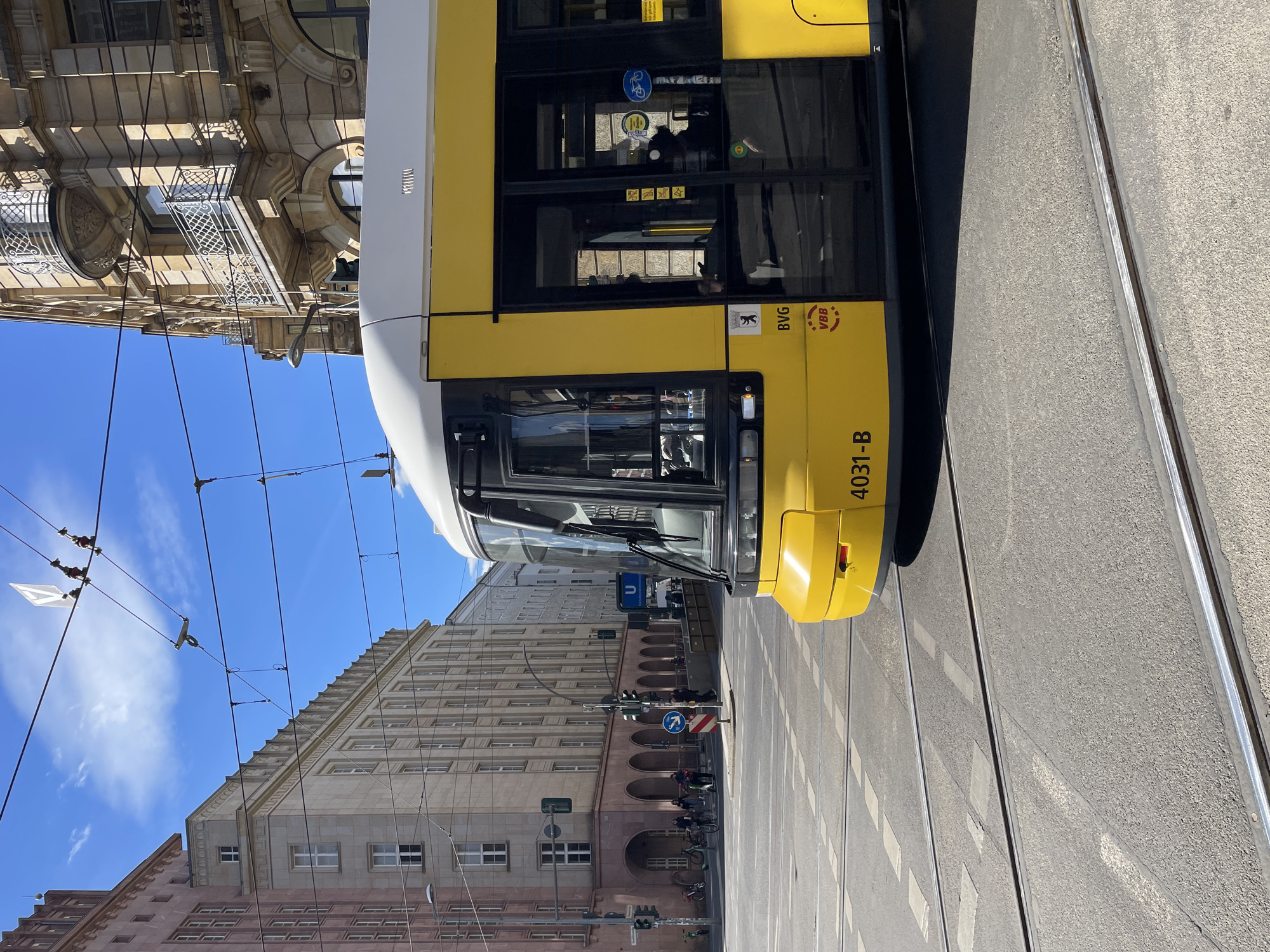 The image captures a bright, sunny urban street scene in Berlin, Germany. A distinctive yellow and white tram, belonging to the Berliner Verkehrsbetriebe (BVG), is the primary subject, occupying the right side of the frame. The tram displays the identification "BVG," "VBB," and the number "4031-B." A bicycle symbol is also visible on one of its windows.
The street features tram tracks running parallel to the tram, with asphalt and road markings visible. Overhead electrical lines for the tram span across a clear, vibrant blue sky dotted with a few white clouds.
On the left side of the street, multi-story buildings with classic European architecture line the thoroughfare. Some buildings exhibit ornate stone facades, decorative balconies, and numerous windows. Further down the street, additional buildings are visible, including one with distinctive ground-floor arches and another with a stepped roofline.
Traffic infrastructure such as traffic lights and street signs are present, including a blue round sign with a white arrow indicating a right turn. A blue "U" sign, denoting an U-Bahn (subway) station, is also visible in the background, confirming the German urban setting.
Several people can be seen inside the tram and in the distance on the street, some appearing to be walking or cycling. Bicycles are parked against a building. The scene depicts a typical daytime moment in a European city, characterized by public transport, historical architecture, and everyday urban activity. The bright sunlight suggests it is midday or early afternoon.