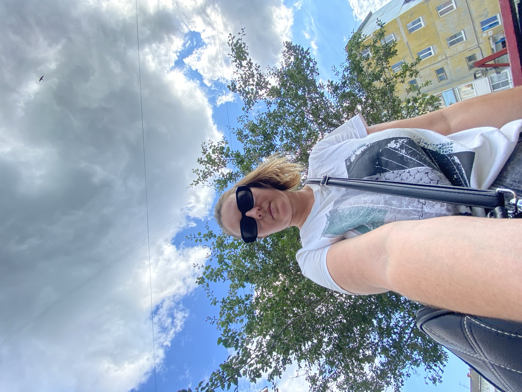 A low-angle, outdoor, medium shot shows a young woman wearing sunglasses and a white t-shirt with a graphic print, holding the camera for a selfie. She is positioned in the foreground, with a lush green tree to her left and a yellow apartment building to her right. The sky above is a mix of blue with white and grey clouds, with power lines crisscrossing the frame. A bird is visible flying in the upper right quadrant of the sky. The scene appears to be in an urban setting, possibly Noviy, Russia, given the architectural style of the building and the presence of power lines.