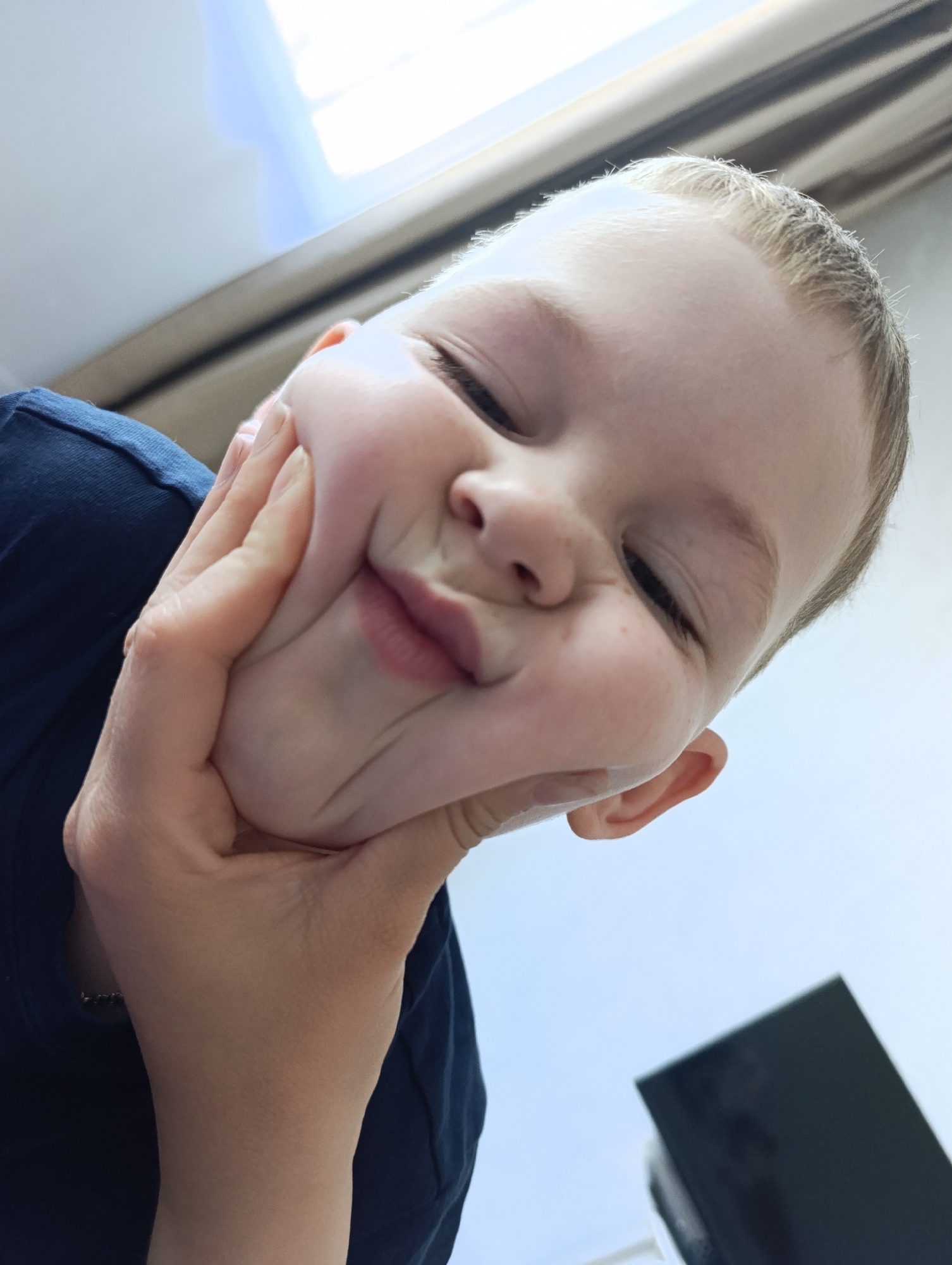A close-up, angled shot depicts a young, light-skinned child with blond hair and closed eyes. The child is holding their inflated cheeks with their right hand, creating a distorted facial expression with pursed lips. They are wearing a dark blue T-shirt. The background includes a light-colored wall or surface and what appears to be a window with drawn curtains. A blurred object, possibly a monitor or electronic device, is visible in the lower right corner. There is no indication of the location being Chaikovsky, Russia, within the visual information provided.