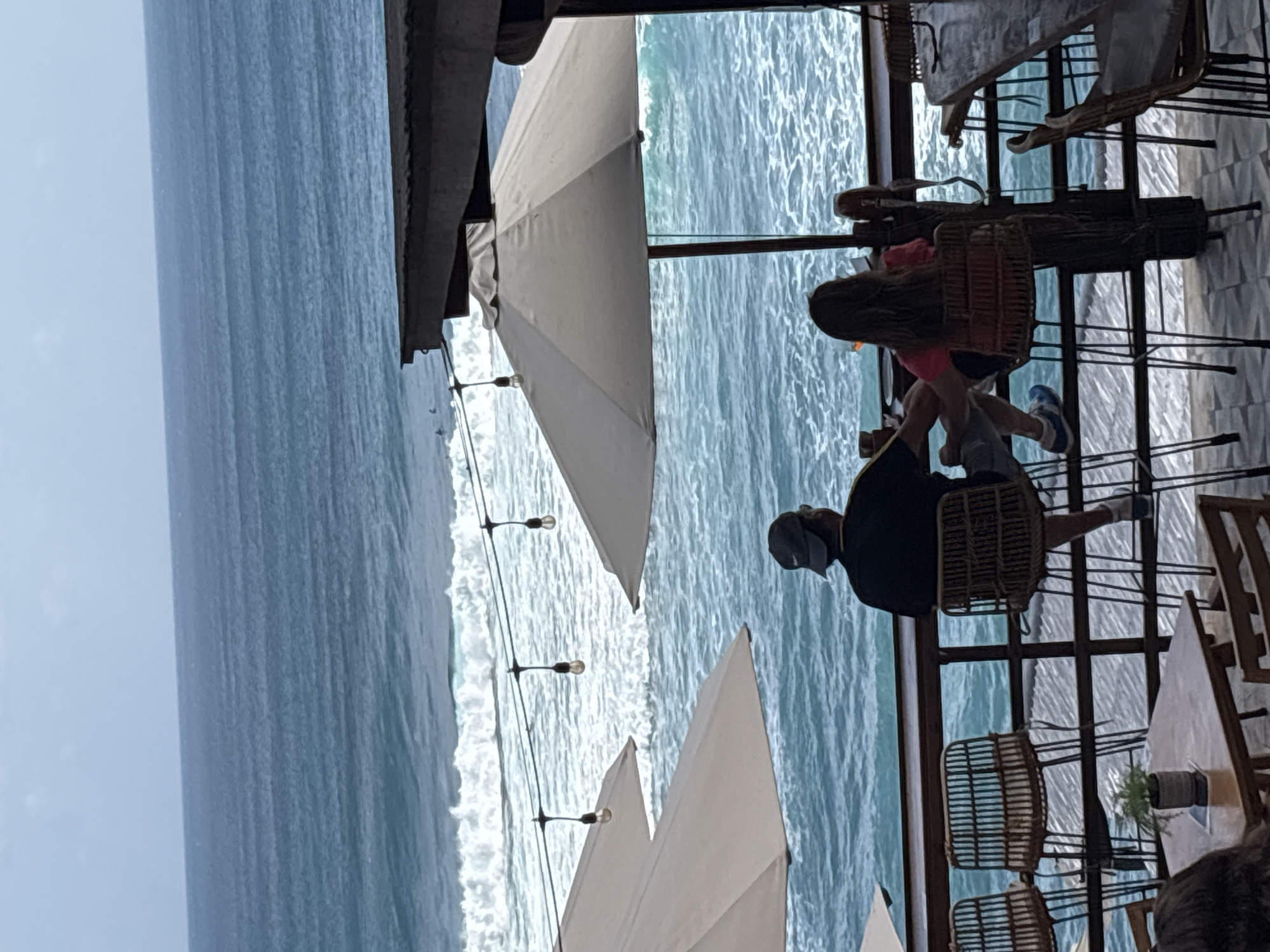 The image captures an elevated outdoor dining or bar area overlooking the ocean in Kuta Selatan, Indonesia. Two individuals are seated with their backs to the viewer at a high-top wooden counter, facing the sea. The person on the left wears a dark t-shirt, a baseball cap, shorts, and blue sneakers, and holds a beverage. The person on the right has long hair, wears a reddish top, and also holds a drink. Both are seated on high-backed rattan stools. In the ocean, white waves are breaking, and multiple small dark figures, identified as surfers, are visible in the water further offshore. The scene includes several large, light-colored patio umbrellas providing shade and a string of exposed light bulbs suspended between the umbrellas and the counter. A dark corrugated roof structure is partially visible on the upper right. Additional rattan seating and a wooden table with a small potted plant are visible in the lower left section of the frame. The sky above the horizon is light blue with minimal cloud cover.